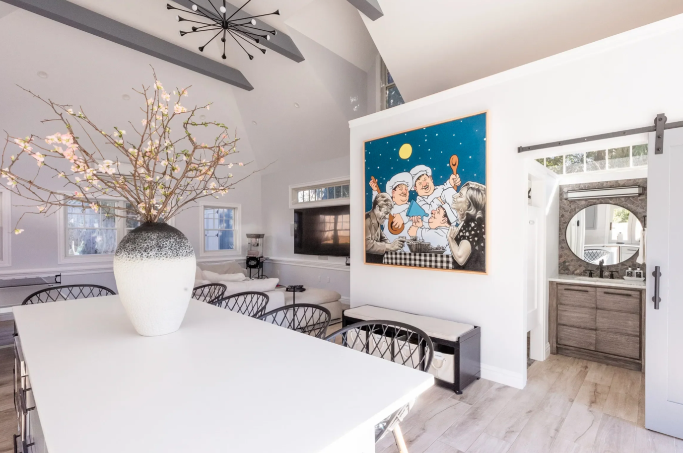 Modern dining room with a large white table, black chairs, a large vase with blossoming branches, and wall art depicting an animated family scene. There are windows, a flat-screen TV, a sliding barn door style closet with a mirror, and a bathroom wit