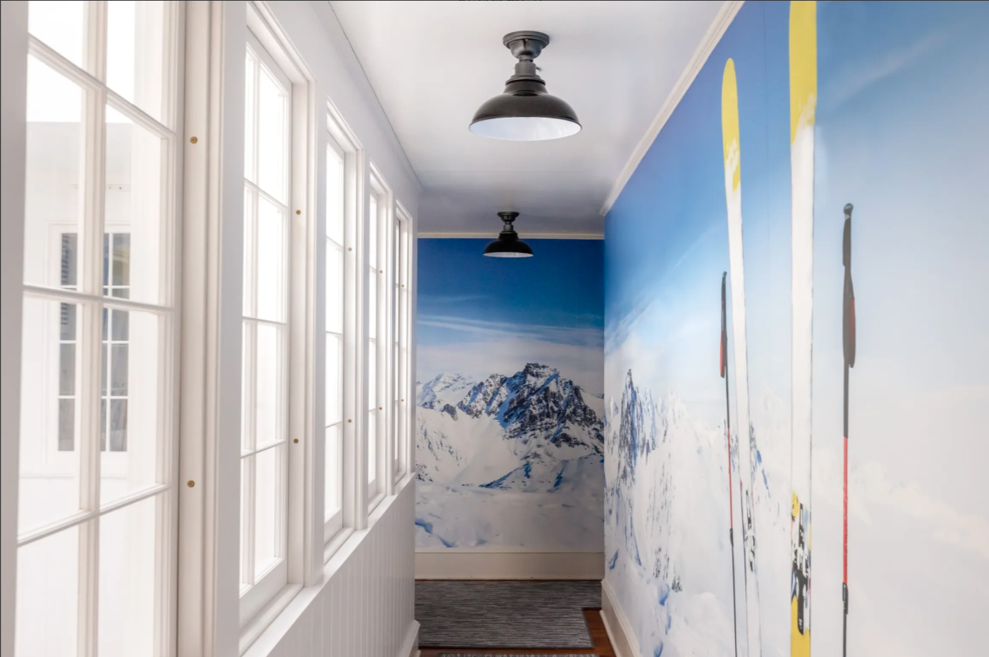 Interior hallway with snow mountain landscape mural on the walls, large windows on the left, and mounted skis displayed on the right.