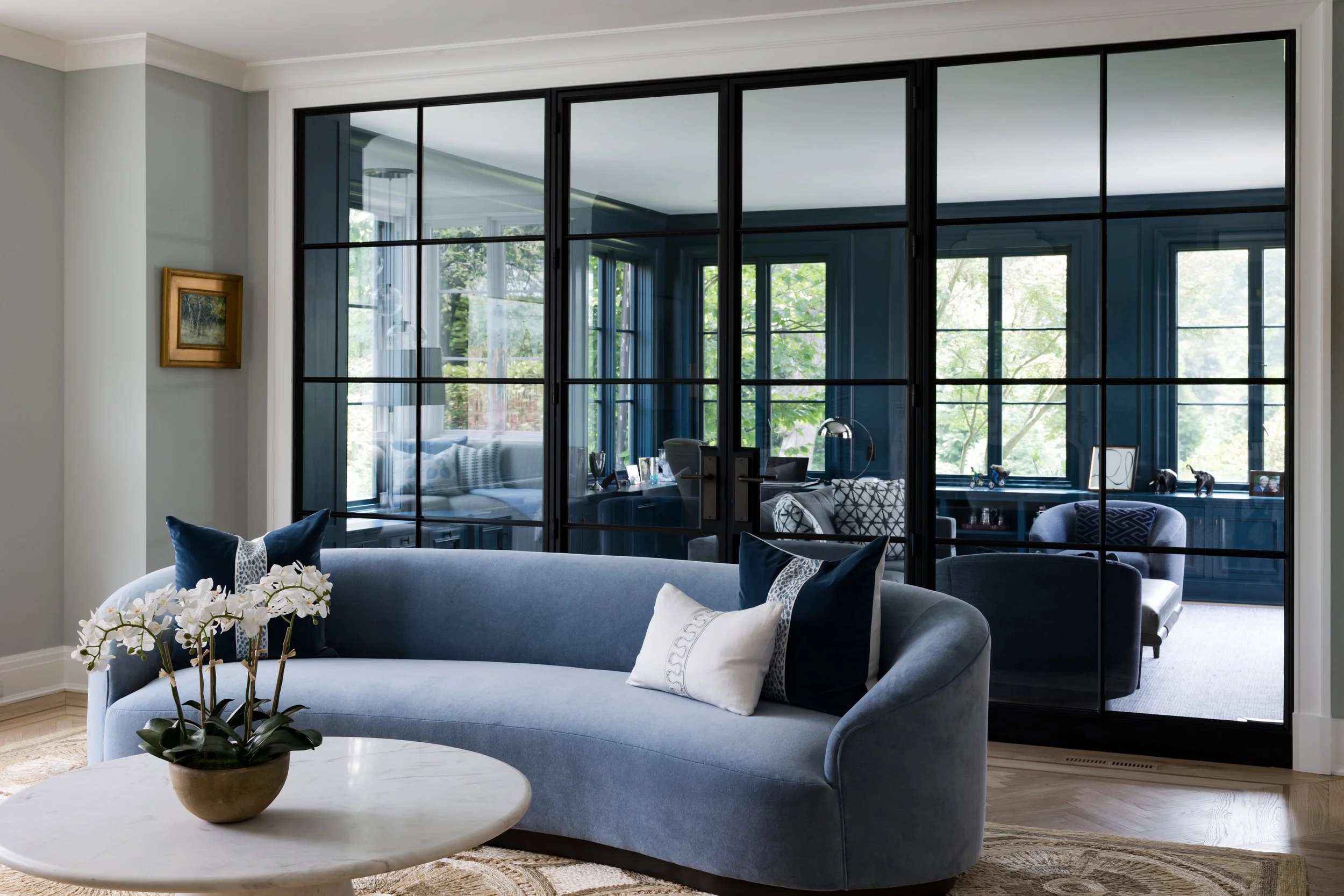Living room with a light blue sofa, decorative pillows, a white round coffee table with a potted orchid, large black framed glass doors, and a view of the lush green outdoors.