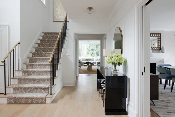 Entryway with staircase on the left, black console table with a flower arrangement and mirror on the right, leading to a living room in the background.
