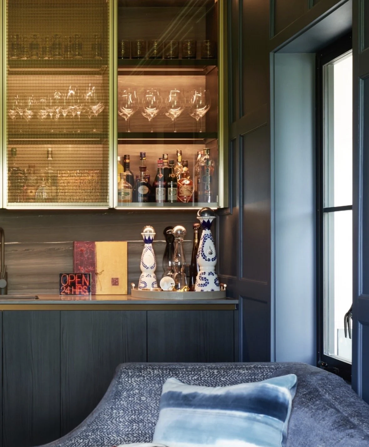 A bar corner with glassware, liquor bottles, decorative vases, a neon "Open 24 Hrs" sign, and a gray sofa with striped pillows beside a window.
