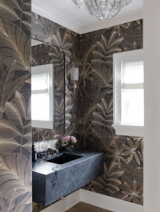 A small bathroom with dark leaf-patterned wallpaper, a black marble countertop sink, a rectangular mirror, a window with white trim, a chandelier on the ceiling, and a wall-mounted light fixture.