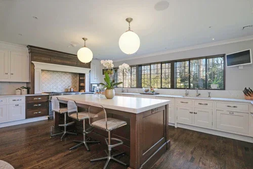 Modern kitchen with white cabinets, a large central island with seating, wooden flooring, and large windows letting in natural light.