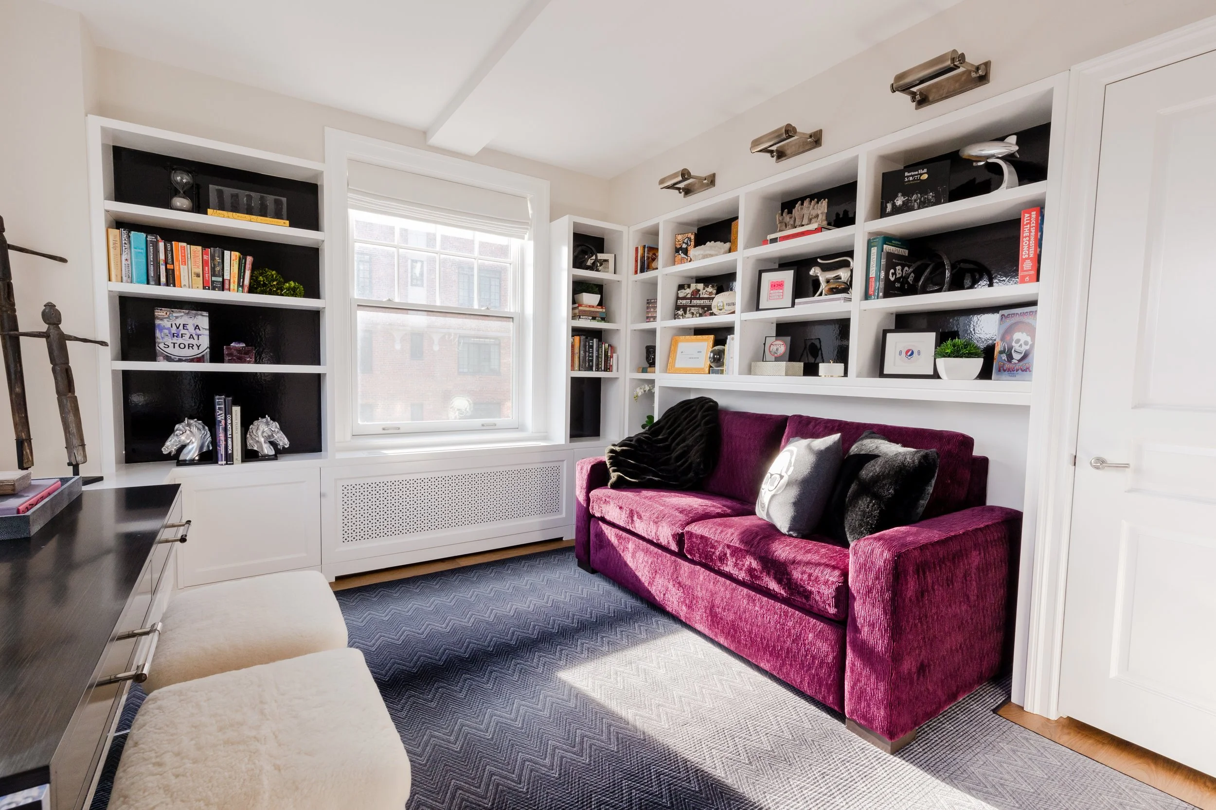 Living room with built-in white shelves filled with books and decor, a window with blinds, a purple velvet sofa with black and white cushions, a light-colored ottoman, patterned gray carpet, and a white door.