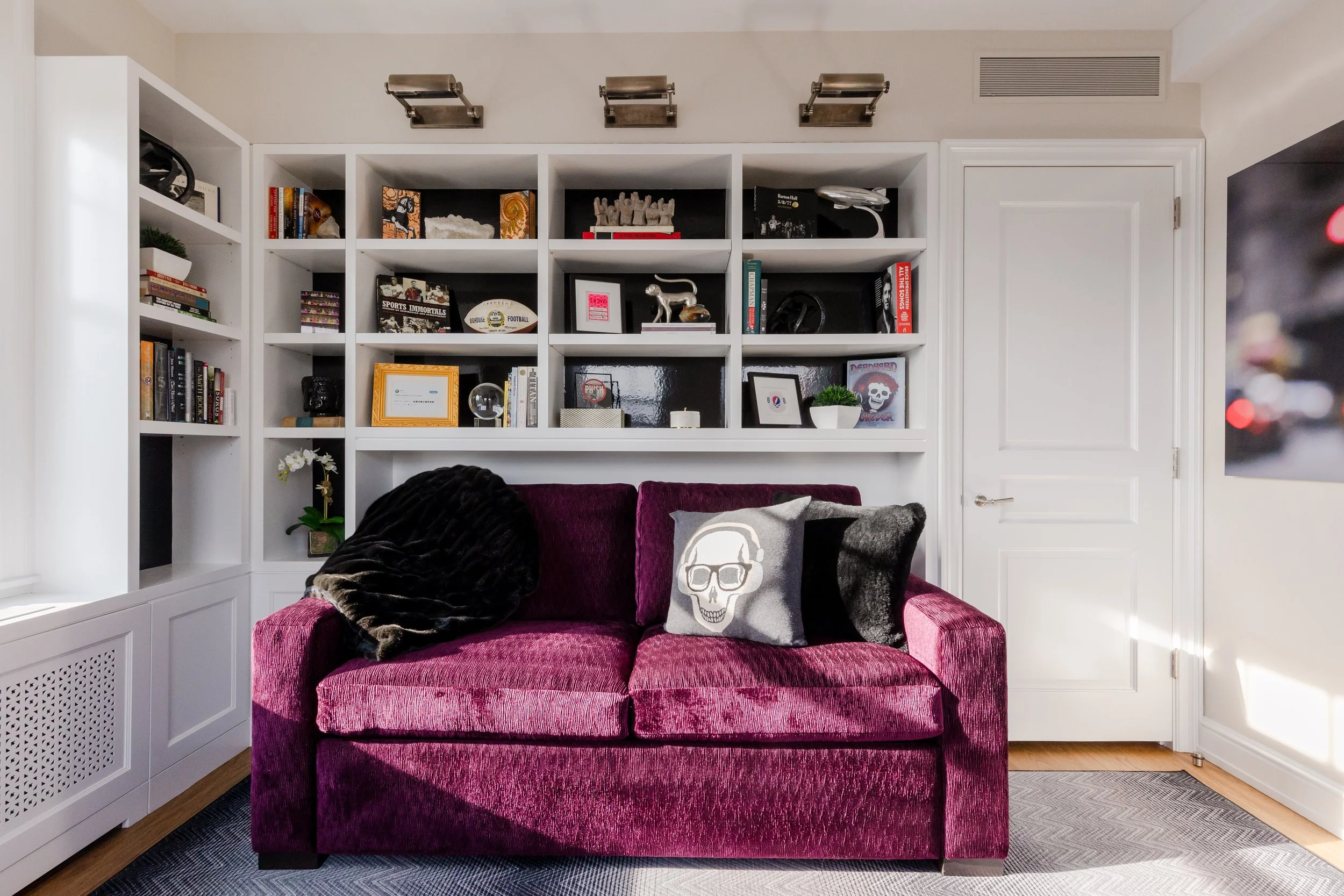 A living room with a magenta velvet armchair with a skull pillow and a draped dark throw blanket, white bookshelf filled with books and decor, and a cream-colored door on the right.