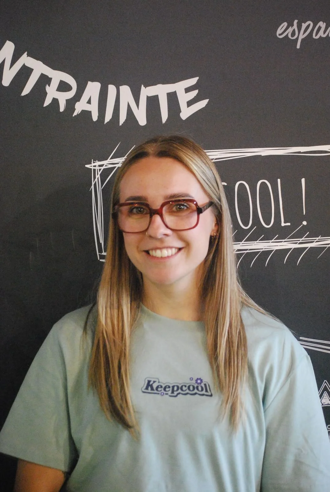 Une jeune femme souriante portant des lunettes rouge foncé et un t-shirt gris avec le logo 'Keepcool' il y a une ardoise noire en arrière-plan avec des mots en français et anglais écrits à la craie blanche.