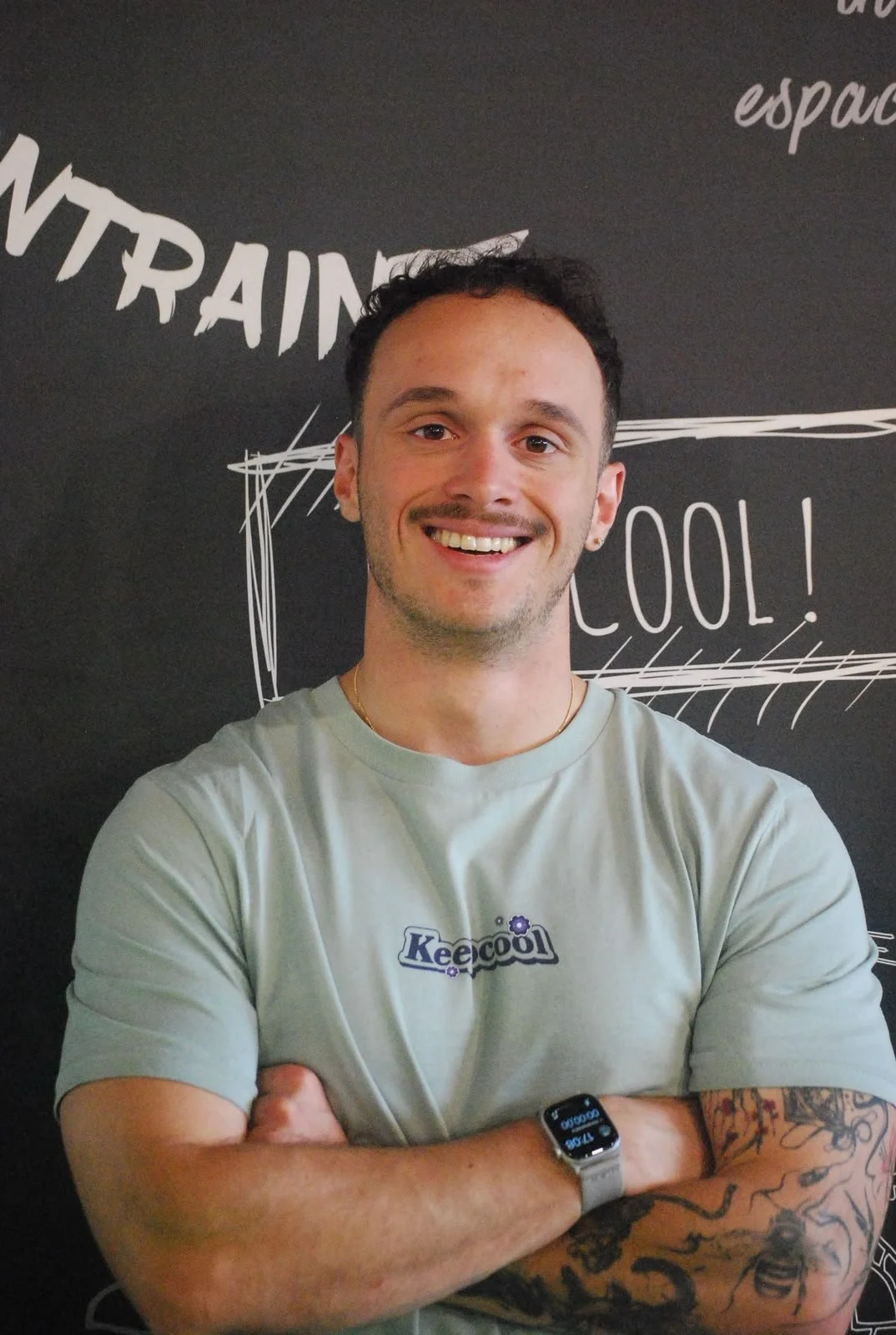 Jeune homme souriant avec cheveux courts, portant un t-shirt gris avec le logo 'Keepcool', montre connectée et bras tatoué, posant devant un mur noir avec écritures blanches.