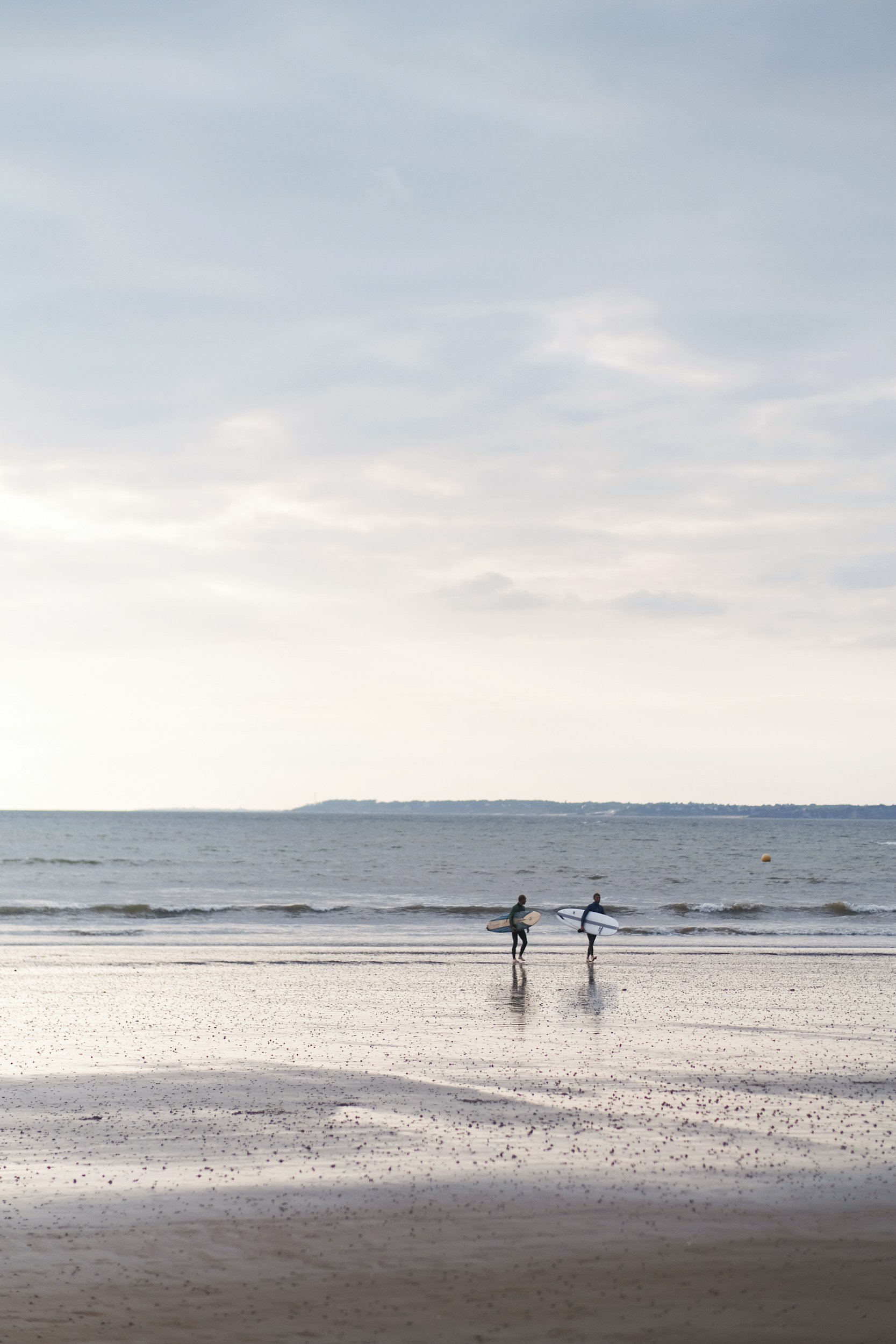 Deux surfeurs marchent vers l'océan avec leurs planches, sous un ciel nuageux, la plage calme en arrière-plan.
