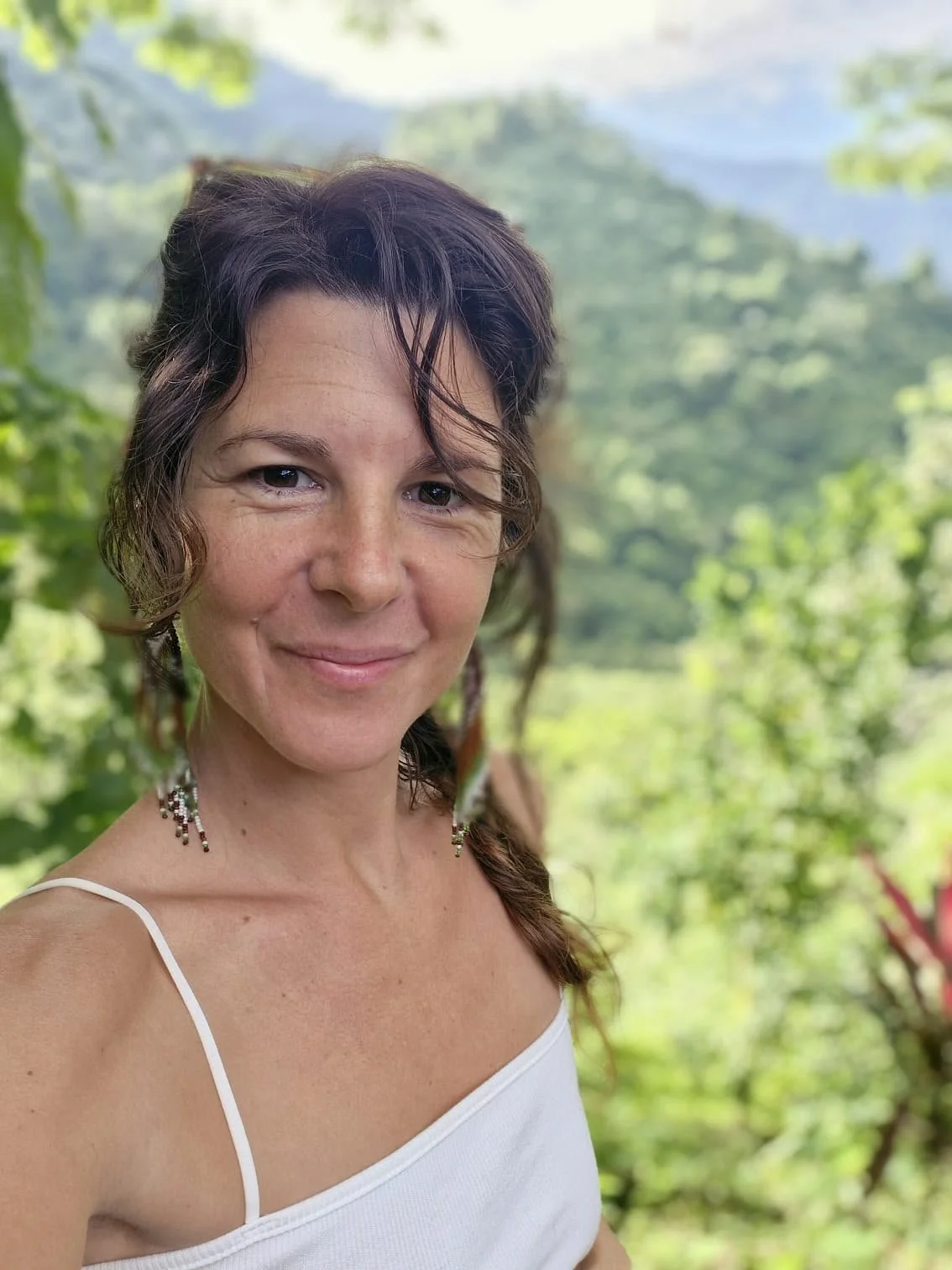 Une femme aux cheveux bruns ondulés et à l'air souriant pose dans un environnement naturel avec des arbres et des montagnes en arrière-plan.