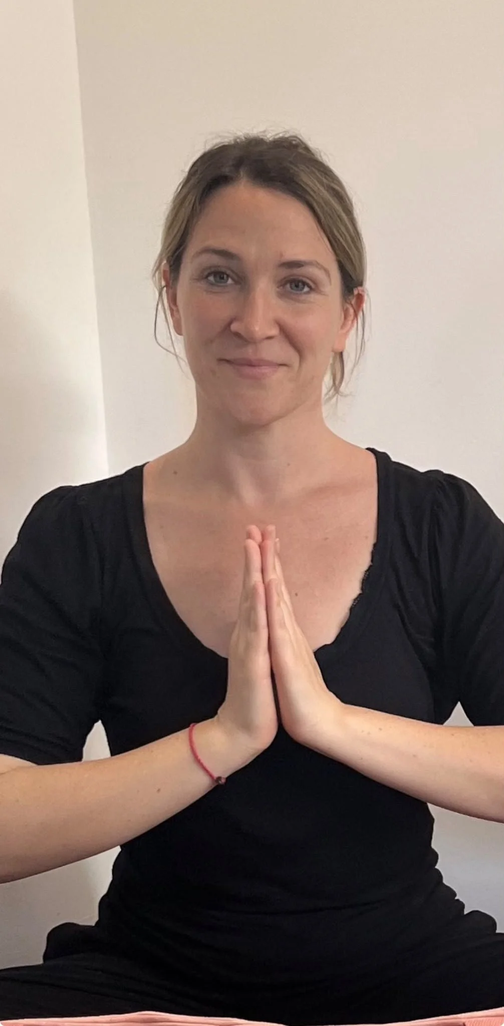 Femme assise en position de méditation ou de yoga, mains jointes en prière, souriant, portant une robe noire, dans une pièce aux murs blancs.