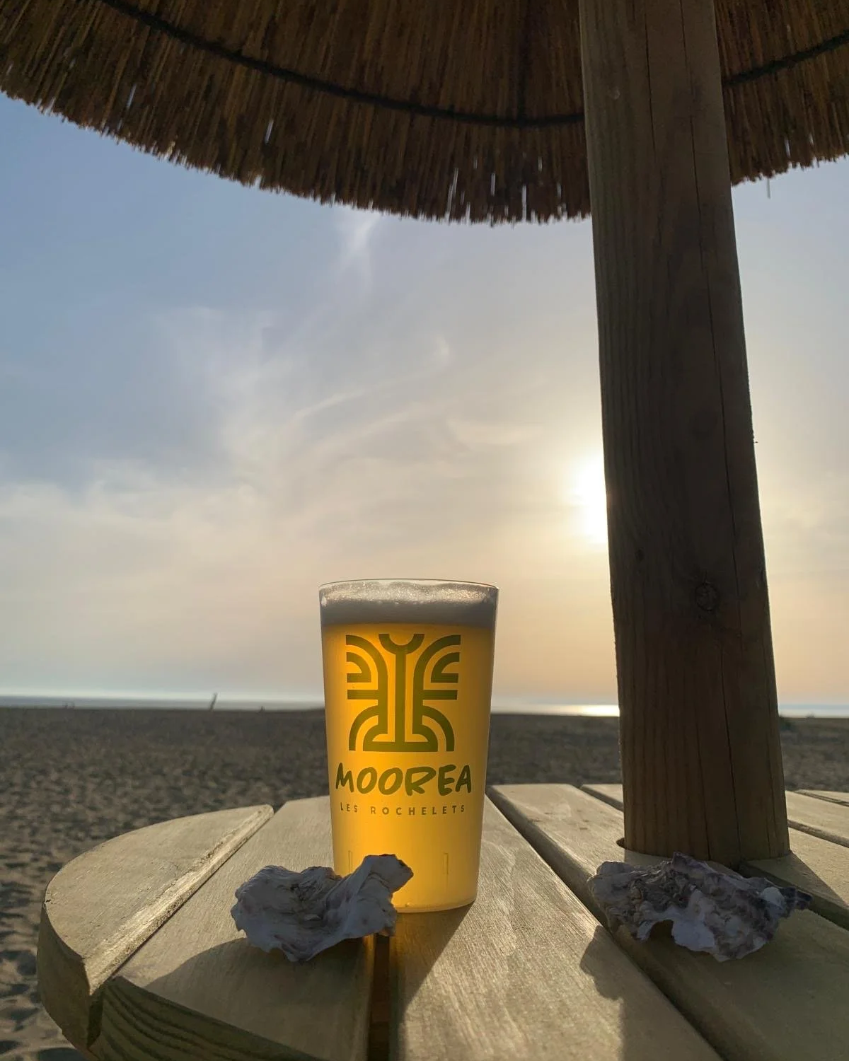 Verre de bière blonde sur une table en bois, sous un parasol en paille, à la plage lors du coucher du soleil.