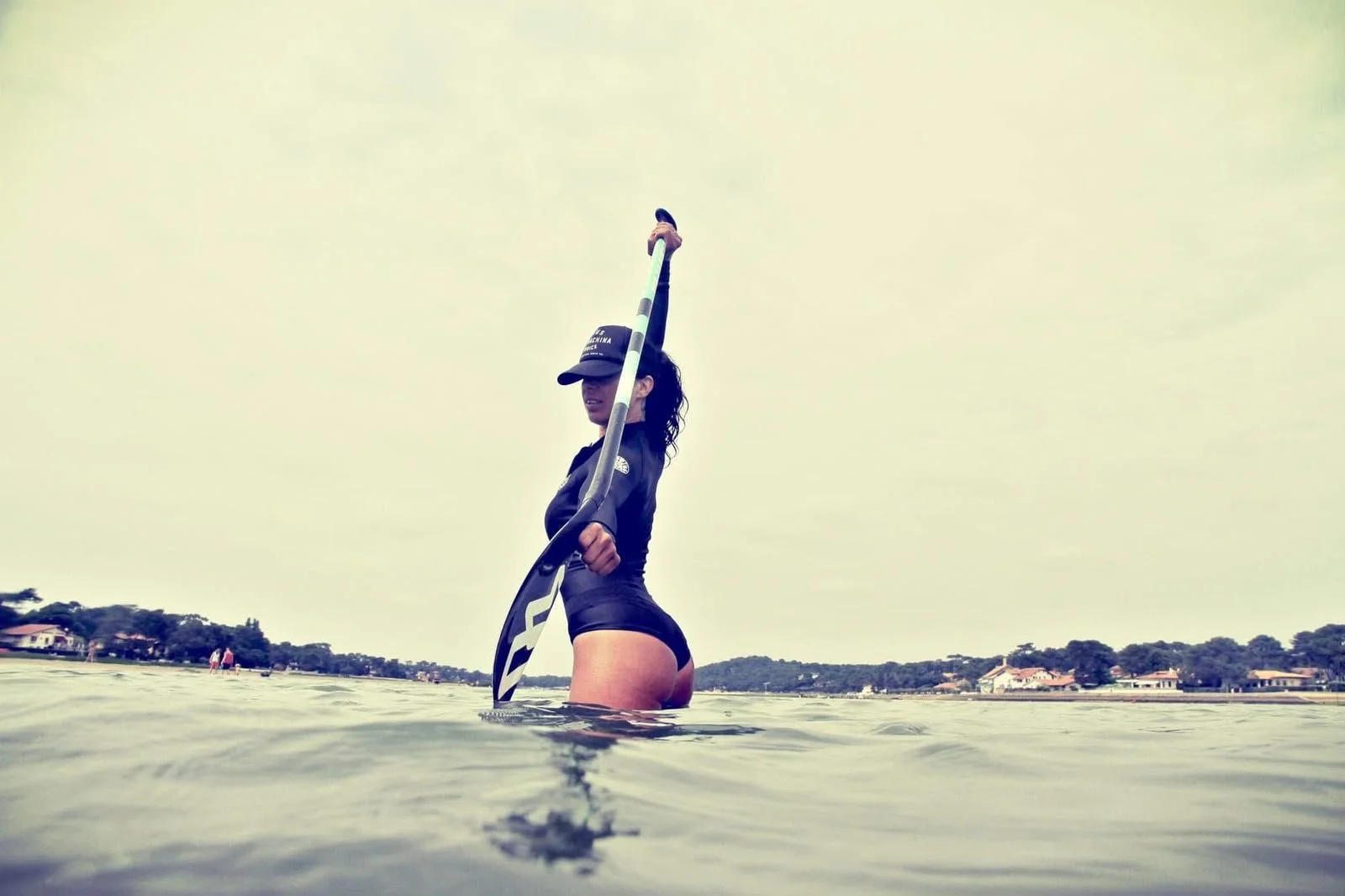 Femme debout dans l'eau, tenant une pagaie, portant une casquette et une tenue de sport, avec un paysage résidentiel en arrière-plan.