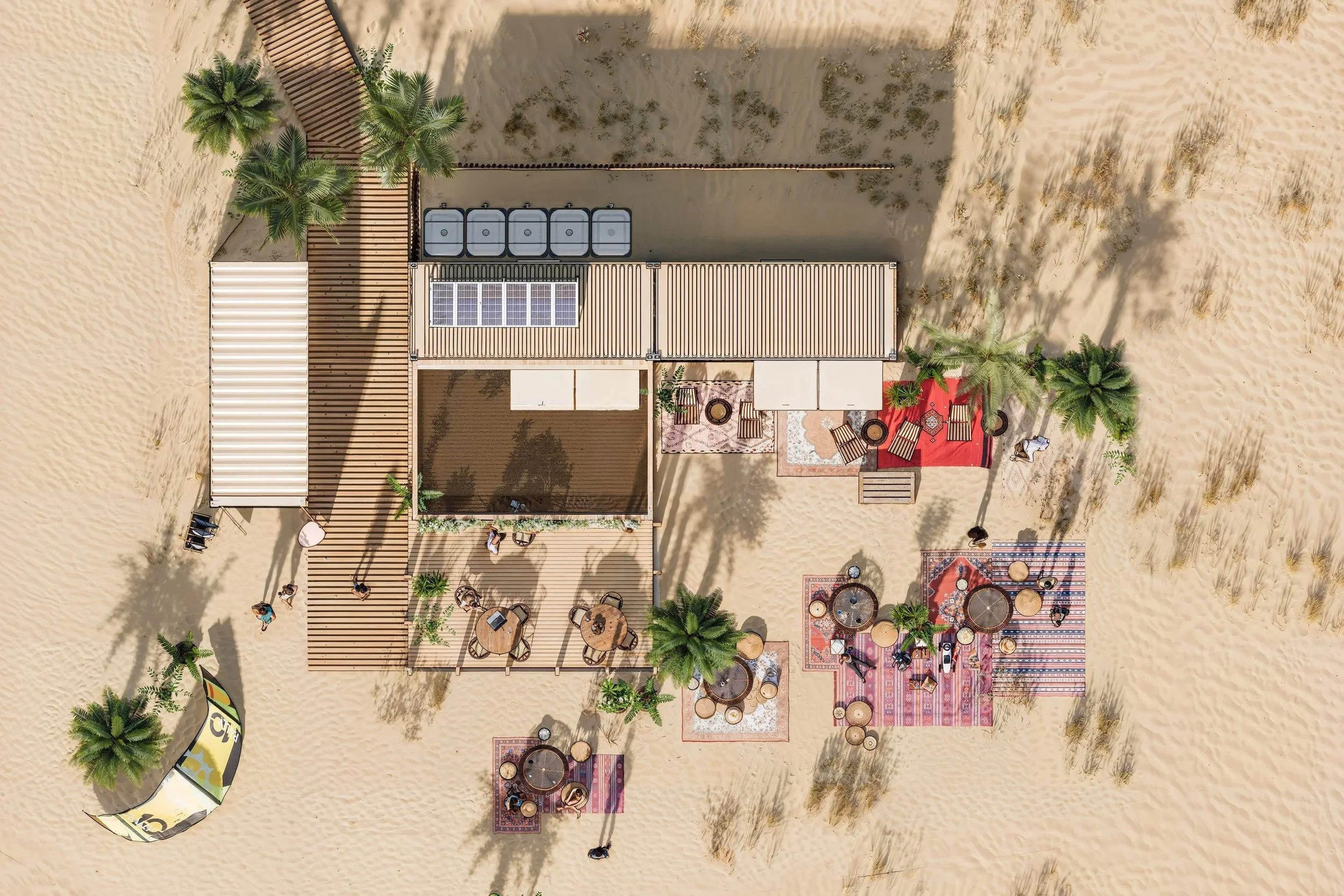 Vue aérienne d'un espace de plage aménagé avec des tables, des fauteuils, des tapis, des parasols, des palmiers et un petit bâtiment en métal. Le tout est entouré par du sable.