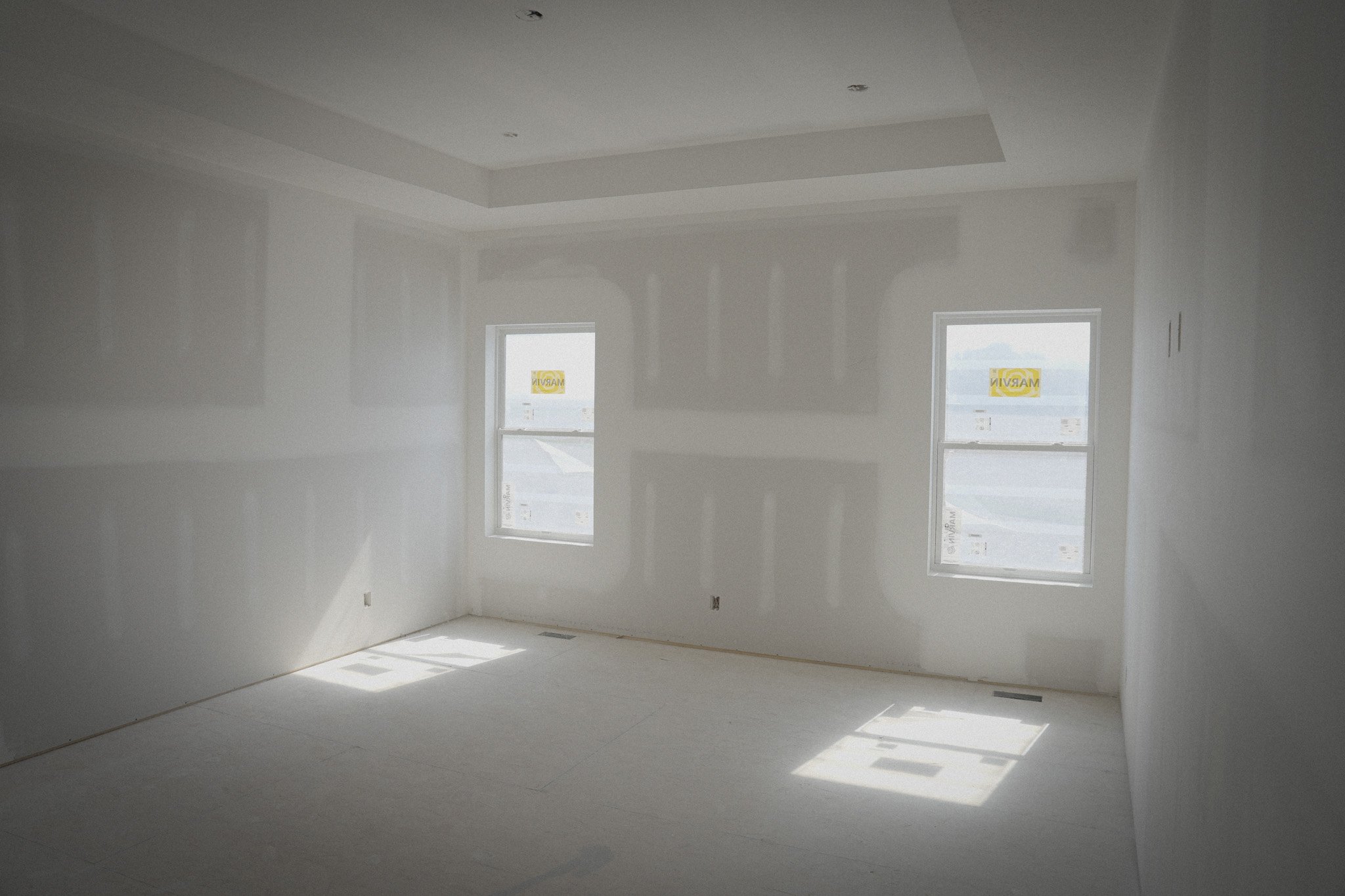 Empty room with white walls, ceiling, and floor, two windows with 'MAPEI' signs, and sunlight casting shadows inside.