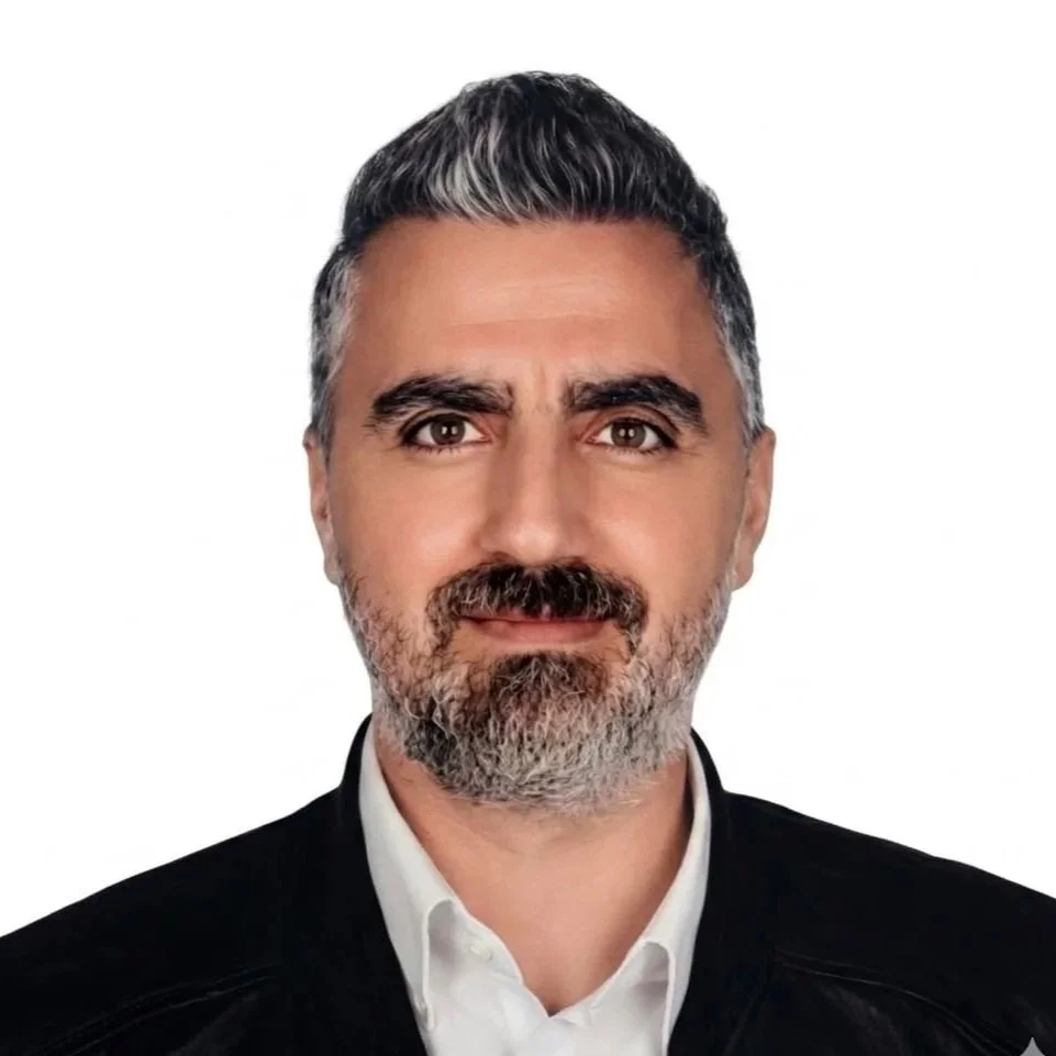 Erkek, gri saçlı, sakallı ve bıyıklı, beyaz gömlek ve siyah ceket giymiş, duruş sergileyen kişi.