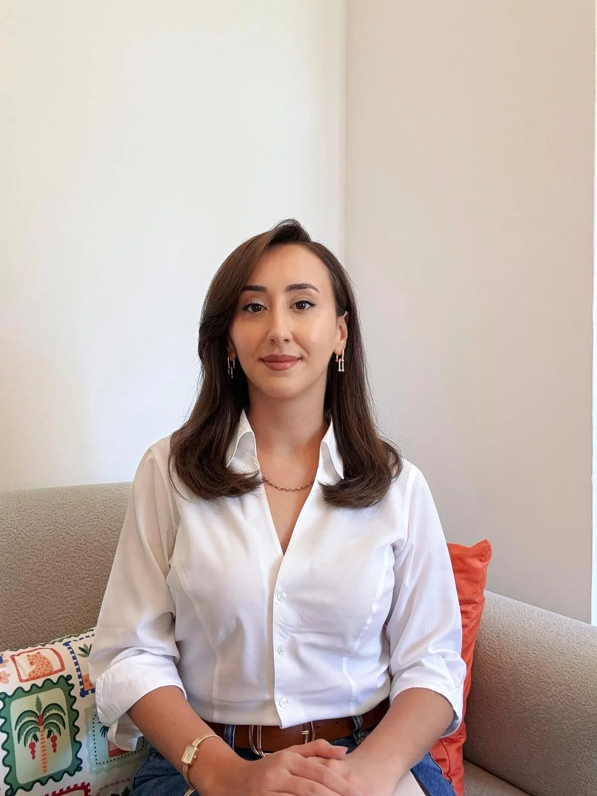 Duvarka giymiş genç kadın, kolundaki saat ve kolyesiyle, rahat bir koltukta oturuyor, arka planda klasik dekor ve pastel duvarlar bulunuyor.