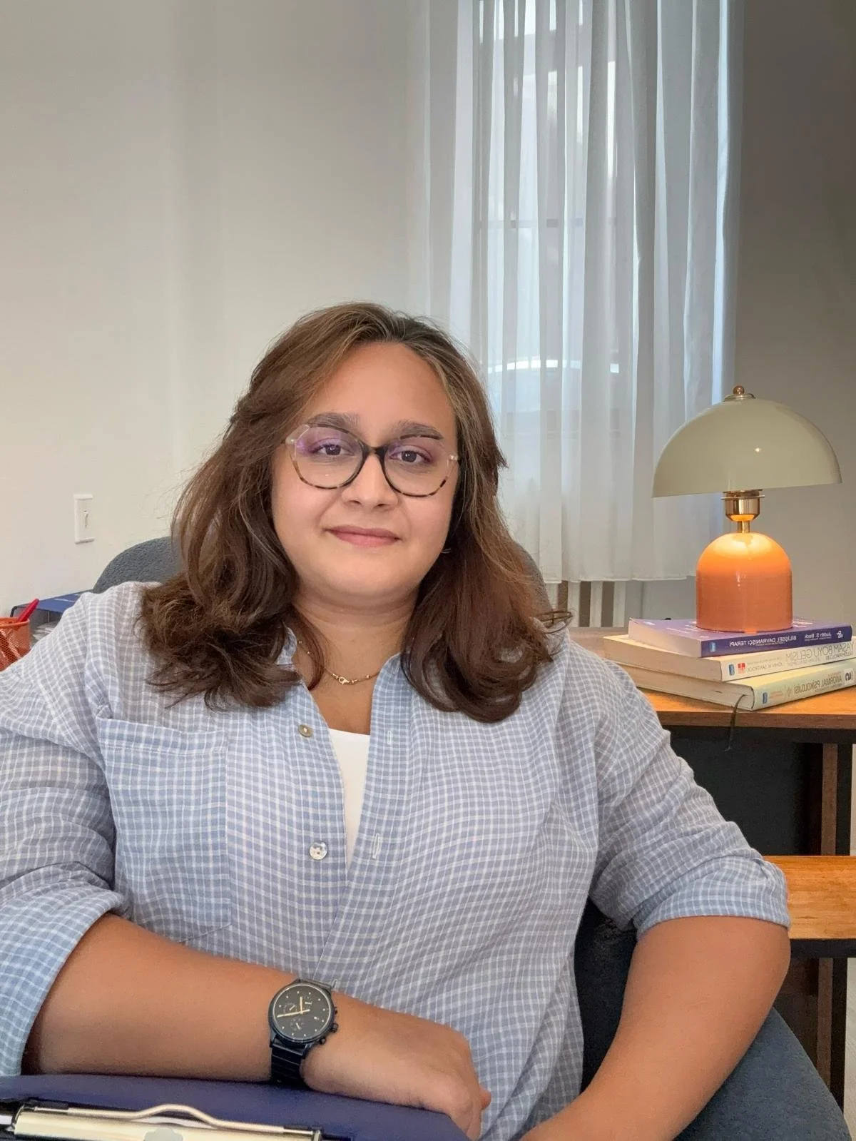 Ofis ortamında oturan kadın. Kadın, kahverengi saçlı, gözlük takıyor ve gülümseyerek bakıyor. Masasında not defteri ve saat var. Arka planda pencere ve masa lambası bulunuyor.