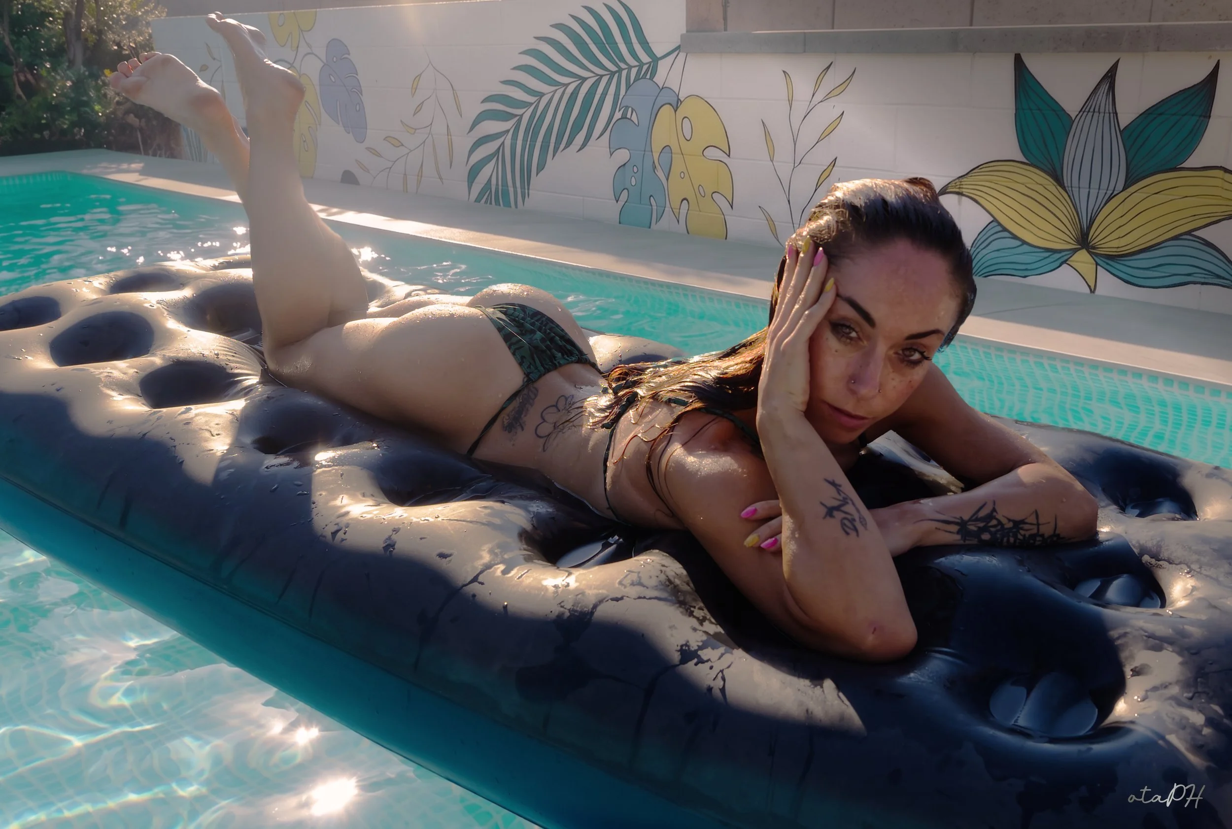Una donna distesa su un materassino nel piscina, con capelli bagnati e che si sfiora la fronte, illuminata dalla luce solare.