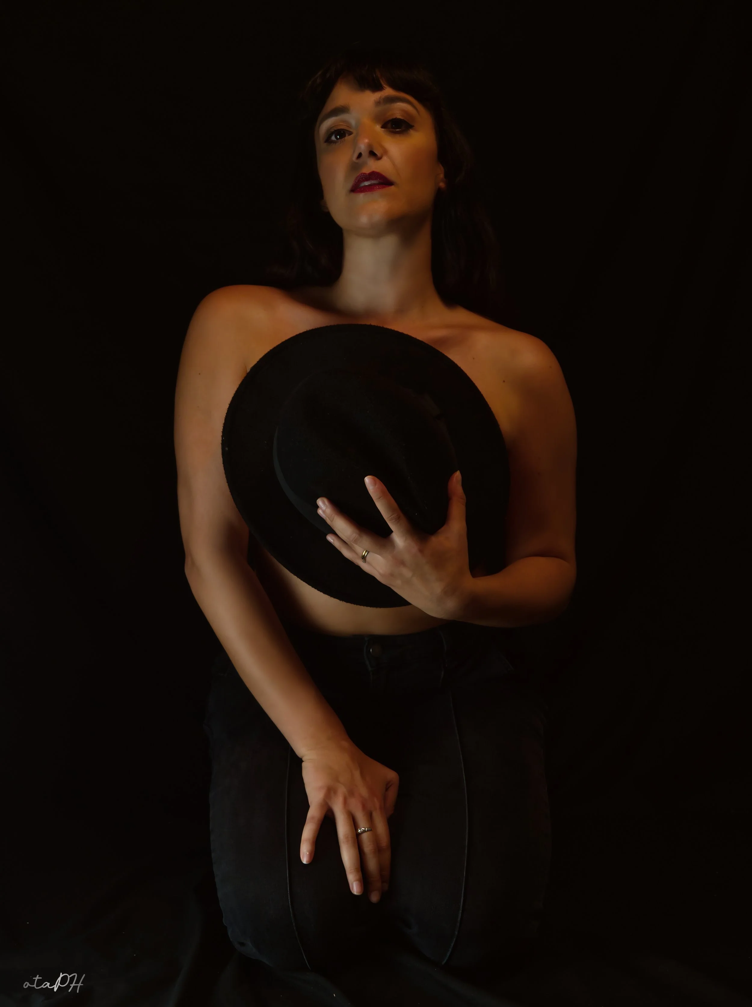 Una donna con i capelli corti e cappelli neri, che tiene un cappello nero davanti al petto, con una mano che fa il gesto di pace e l'altra che tocca la gamba, sotto uno sfondo nero.