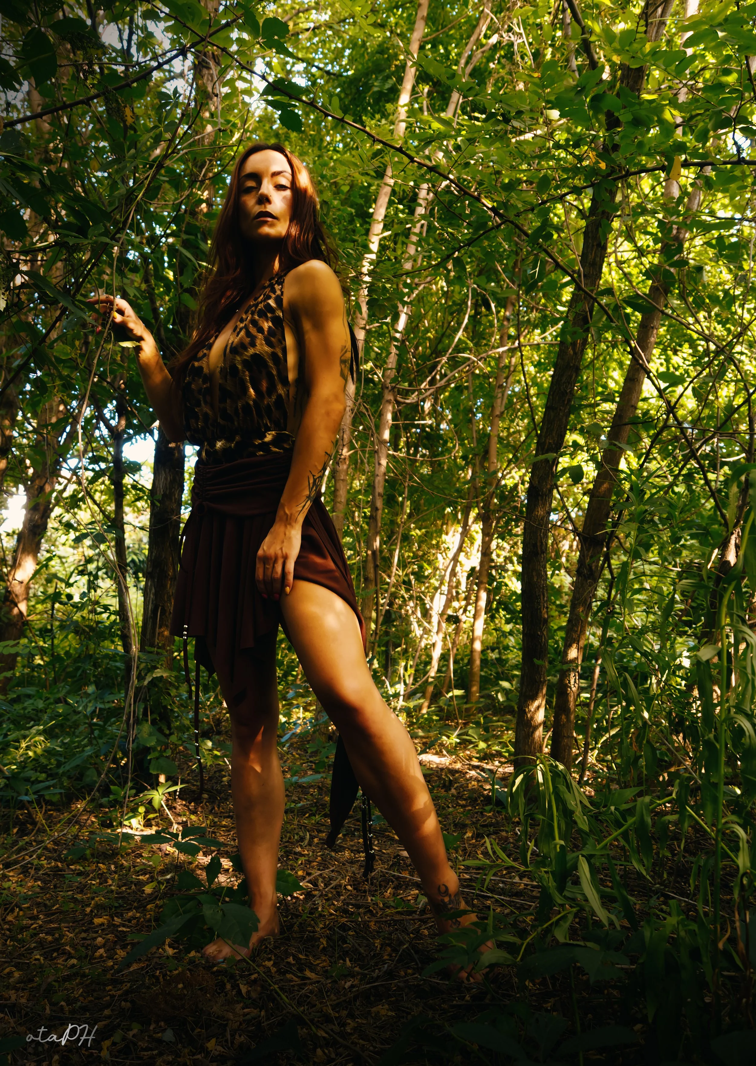 Una donna in abiti leopardati e una gonna lunga, senza scarpe, si trova in una foresta con molti alberi e foglie verdi. La luce del sole filtra attraverso le rami, creando un'atmosfera misteriosa e naturale.