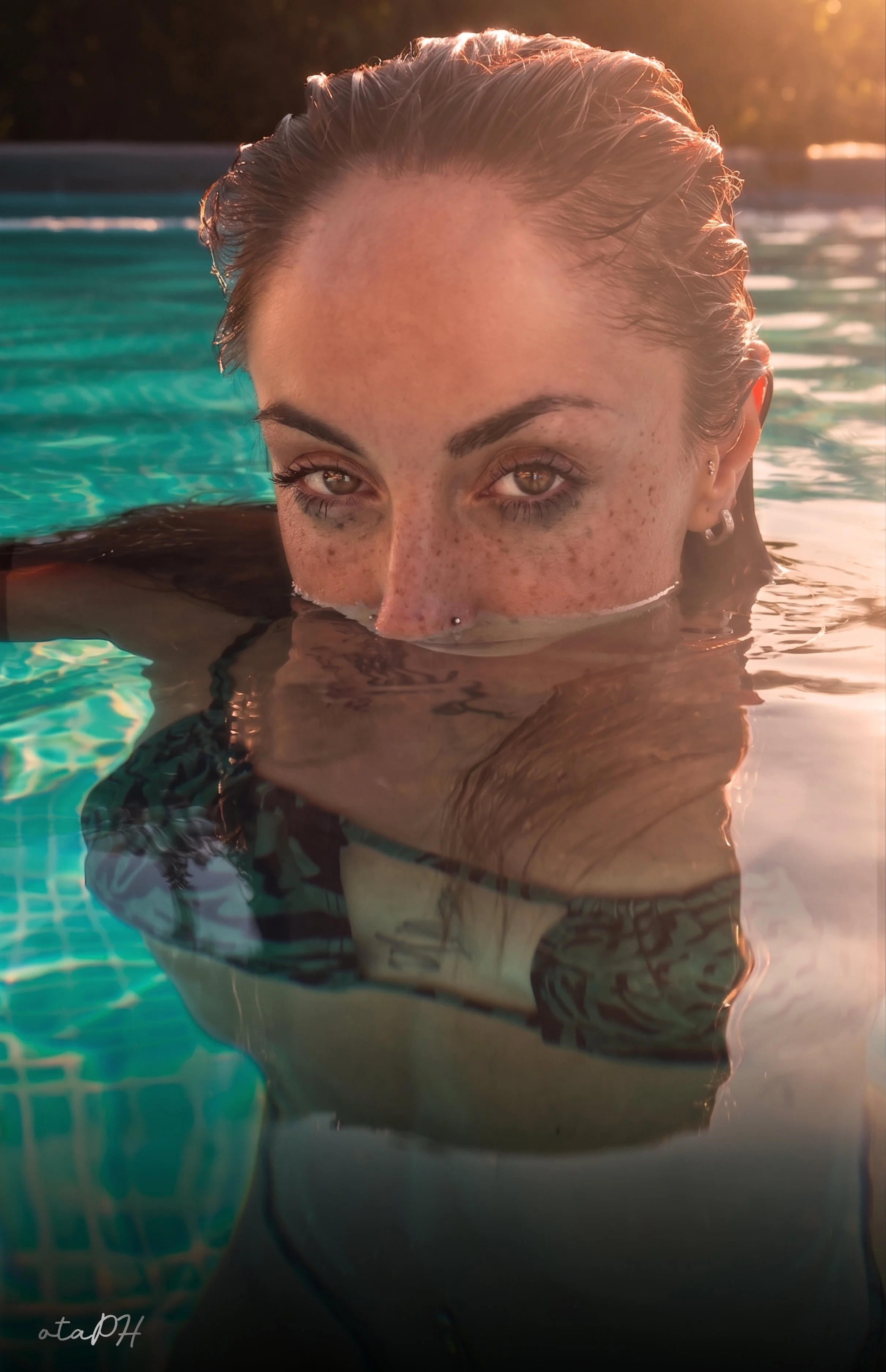 Ragazza con lentiggini che nuota in piscina con acqua cristallina, luce solare calda sul viso, capelli bagnati, incrocia lo sguardo con la fotocamera.