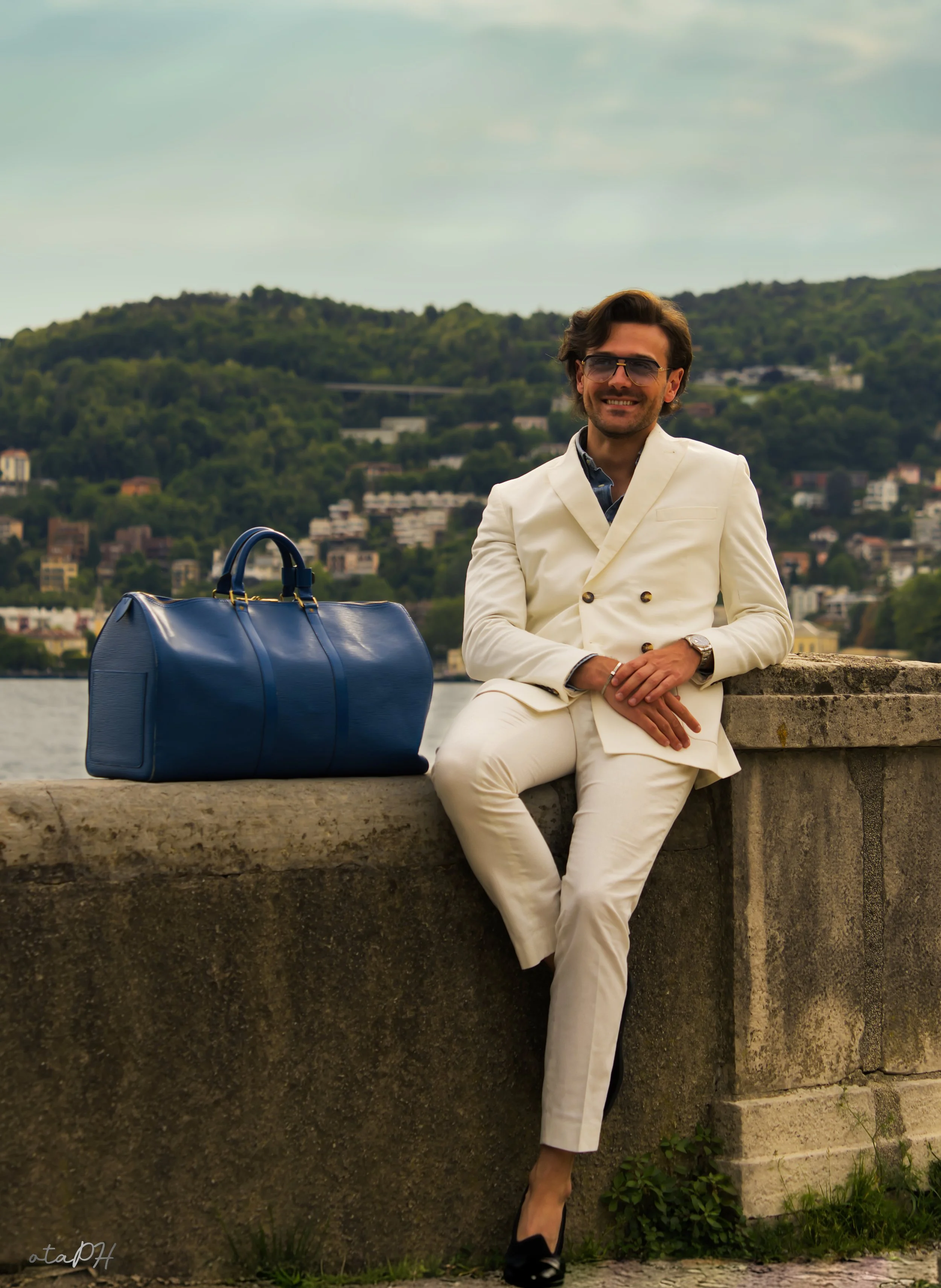 Uomo vestito elegantemente in abito bianco, seduto su un muretto di pietra vicino a un fiume con un paesaggio di colline verdi sullo sfondo. Ha occhiali da sole e una grande borsa blu accanto a lui.