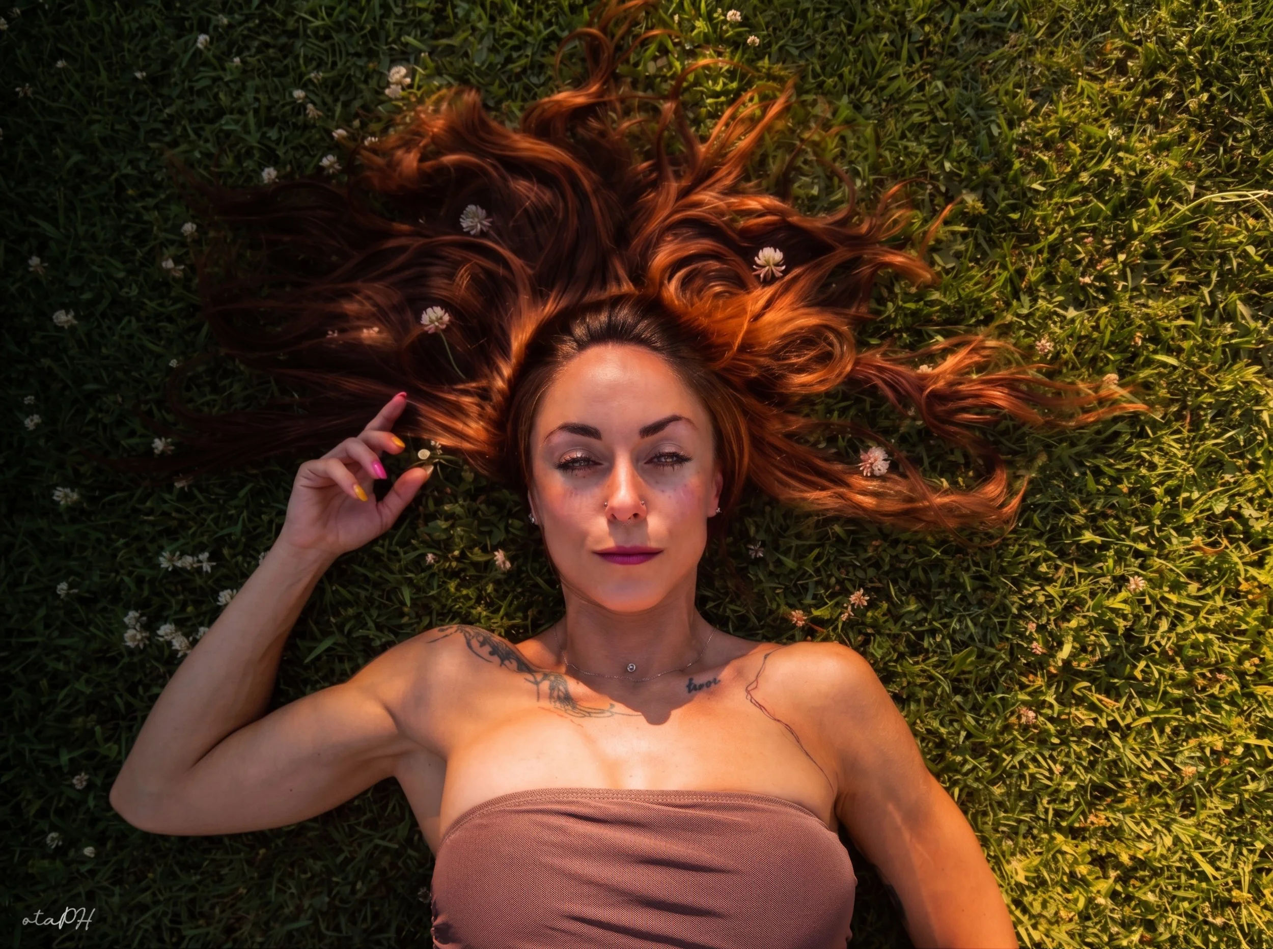 Una donna sdraiata sull'erba con lunghe ciocche di capelli rossi pieni di fiori bianchi, occhi chiusi, tatuaggi sul braccio e sul petto, indossa un abbigliamento color carne e tiene una mano vicino al volto.