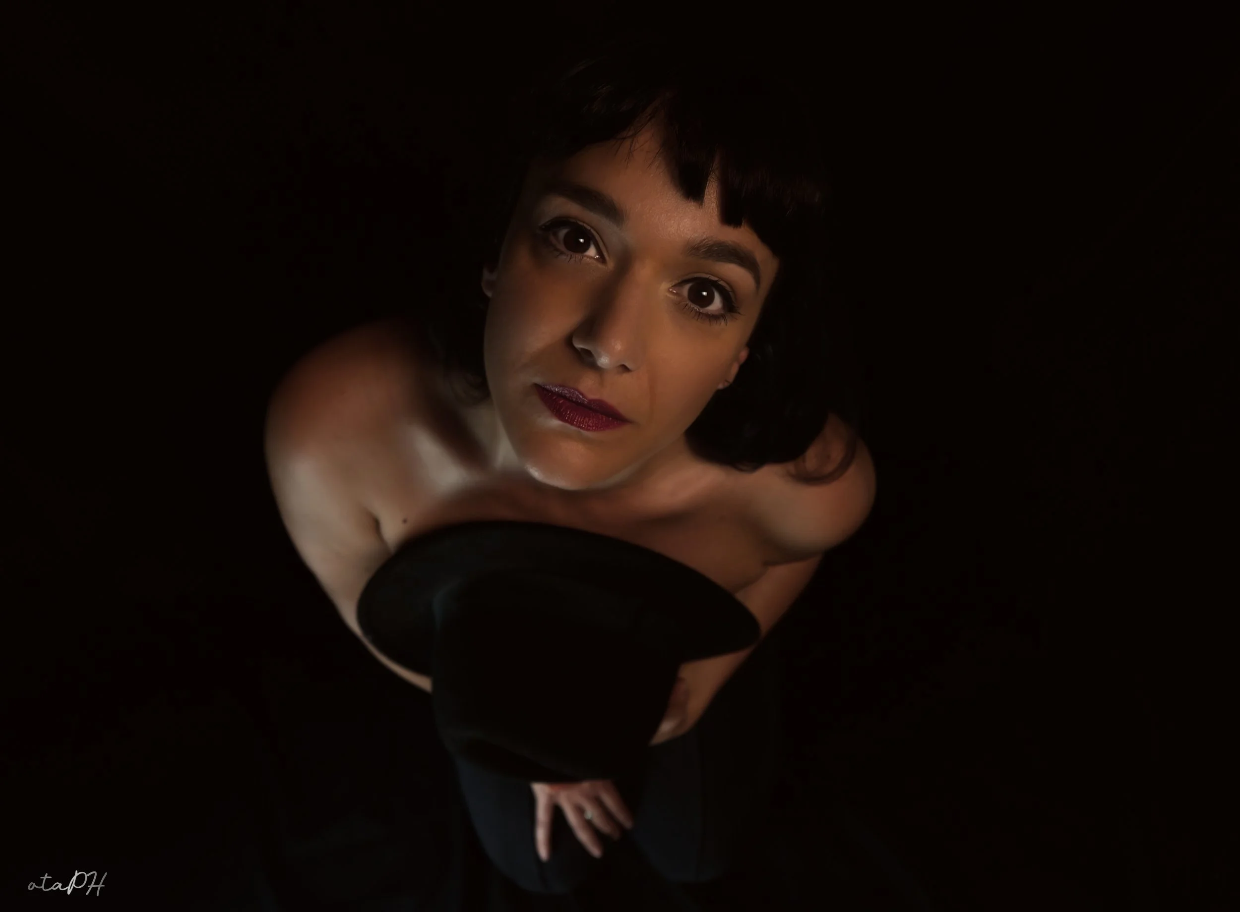 Ritratto di una donna con capelli corti e occhi scuri, che indossa guanti neri, posizionata in modo suggestivo contro uno sfondo nero.