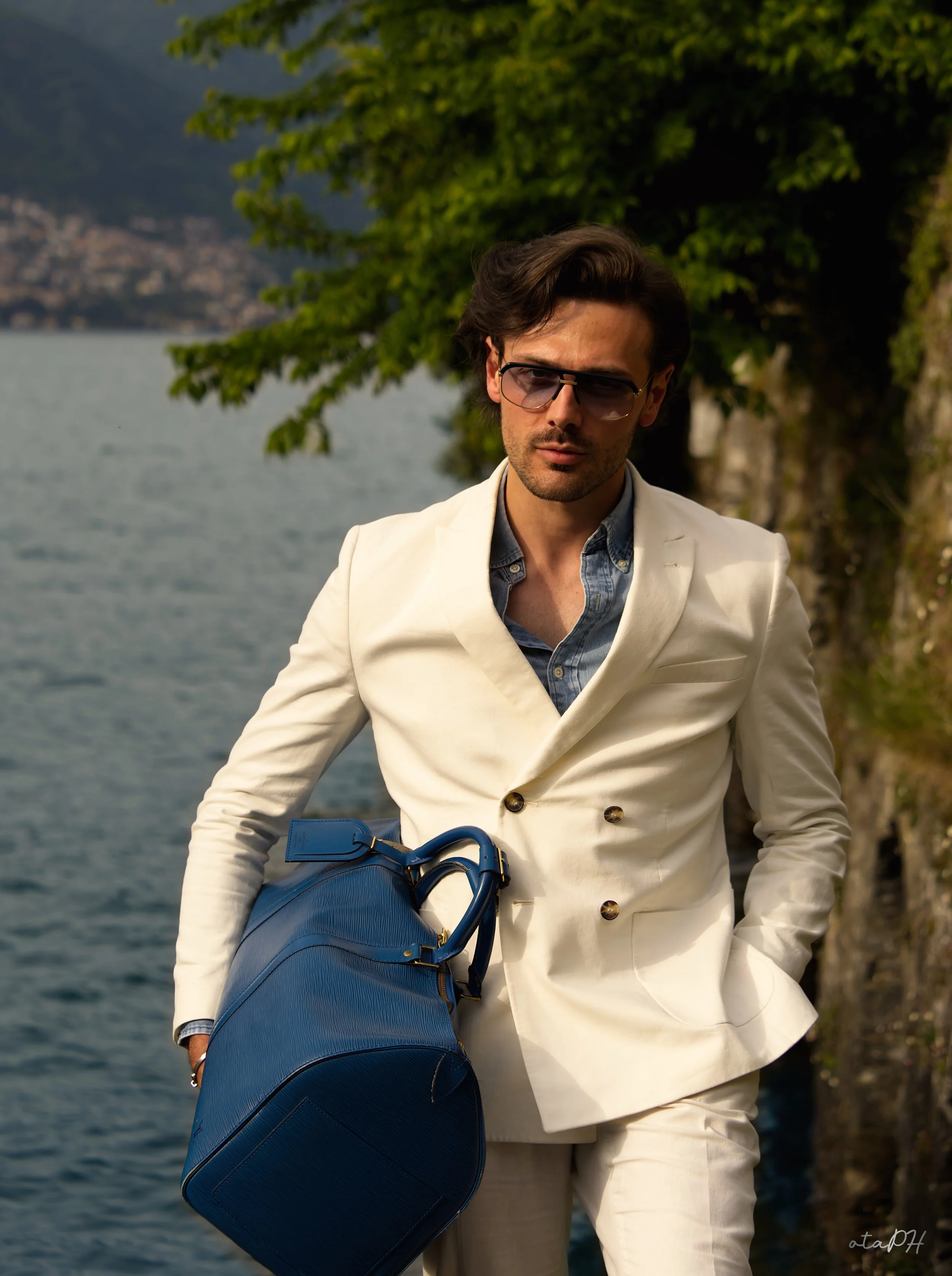 Uomo con occhiali da sole, vestito elegante beige, porta una borsa blu e si trova vicino a un lago con alberi sullo sfondo.