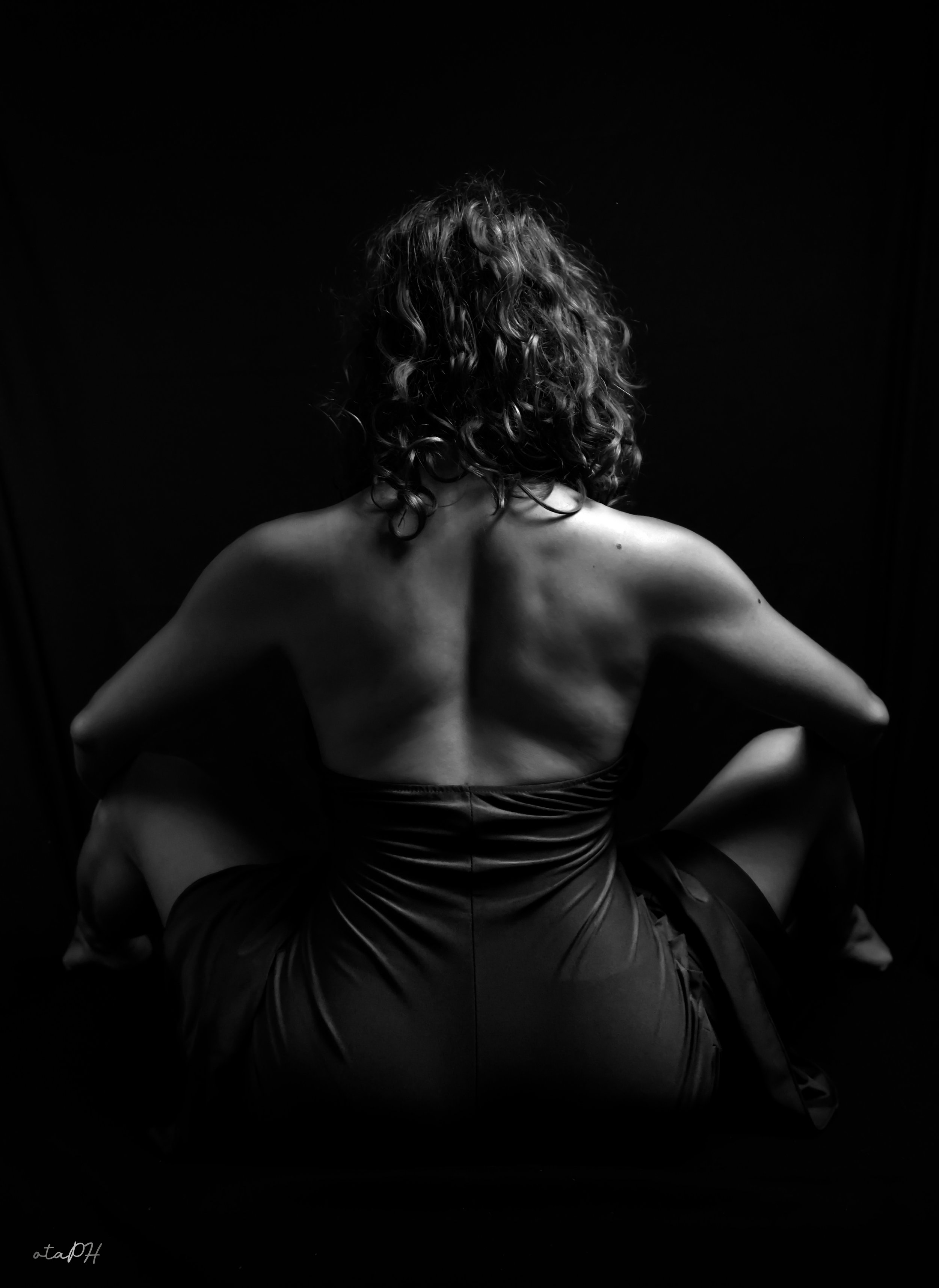 Una donna con i capelli ricci e lunghi vista da dietro, seduta con le gambe incrociate, abbigliamento in satin scuro, sfondo nero.