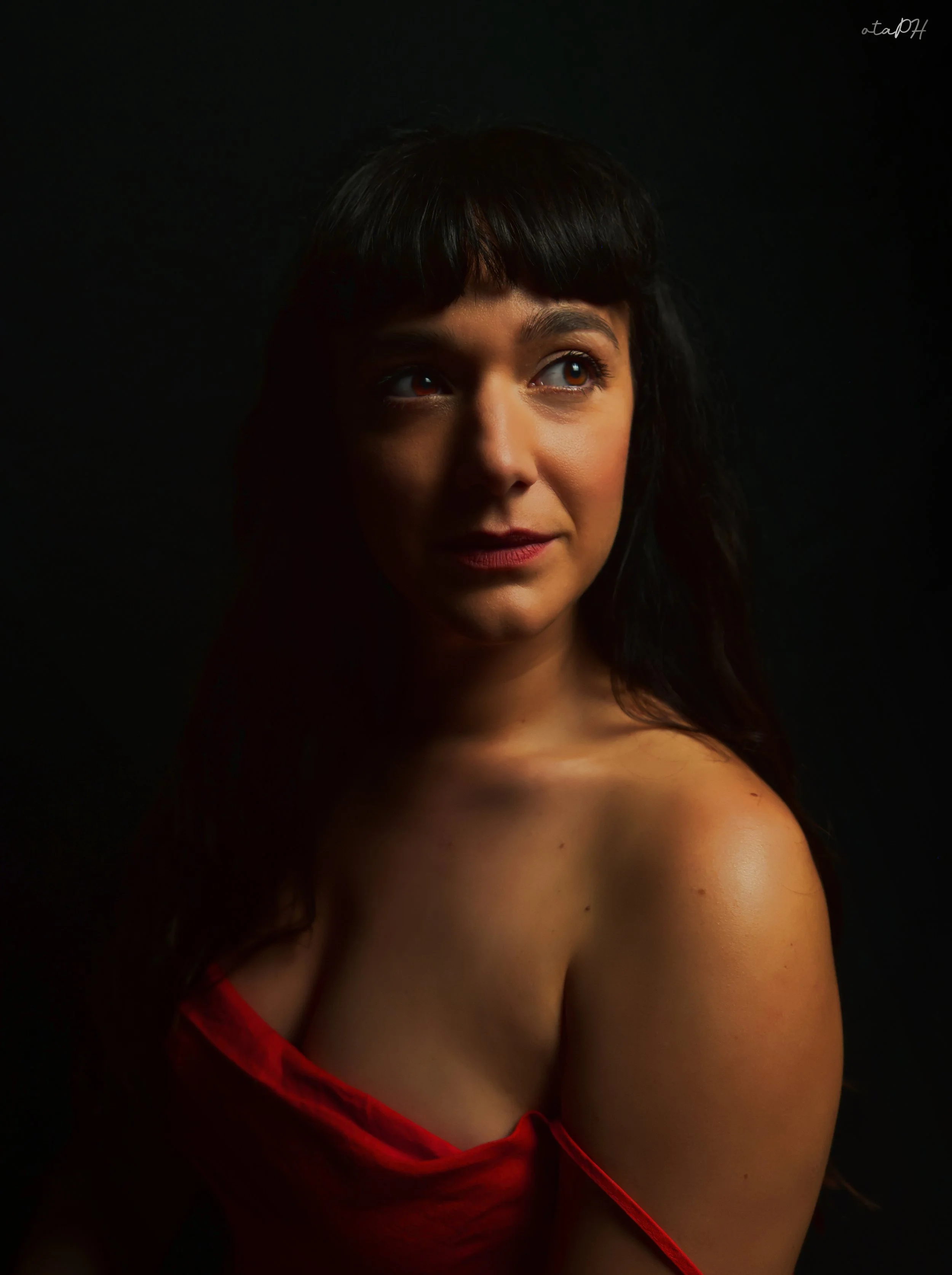 Ritratto di una giovane donna con capelli scuri, occhi castani e espressione sorridente, indossa un vestito rosso in un ambiente con sfondo nero.
