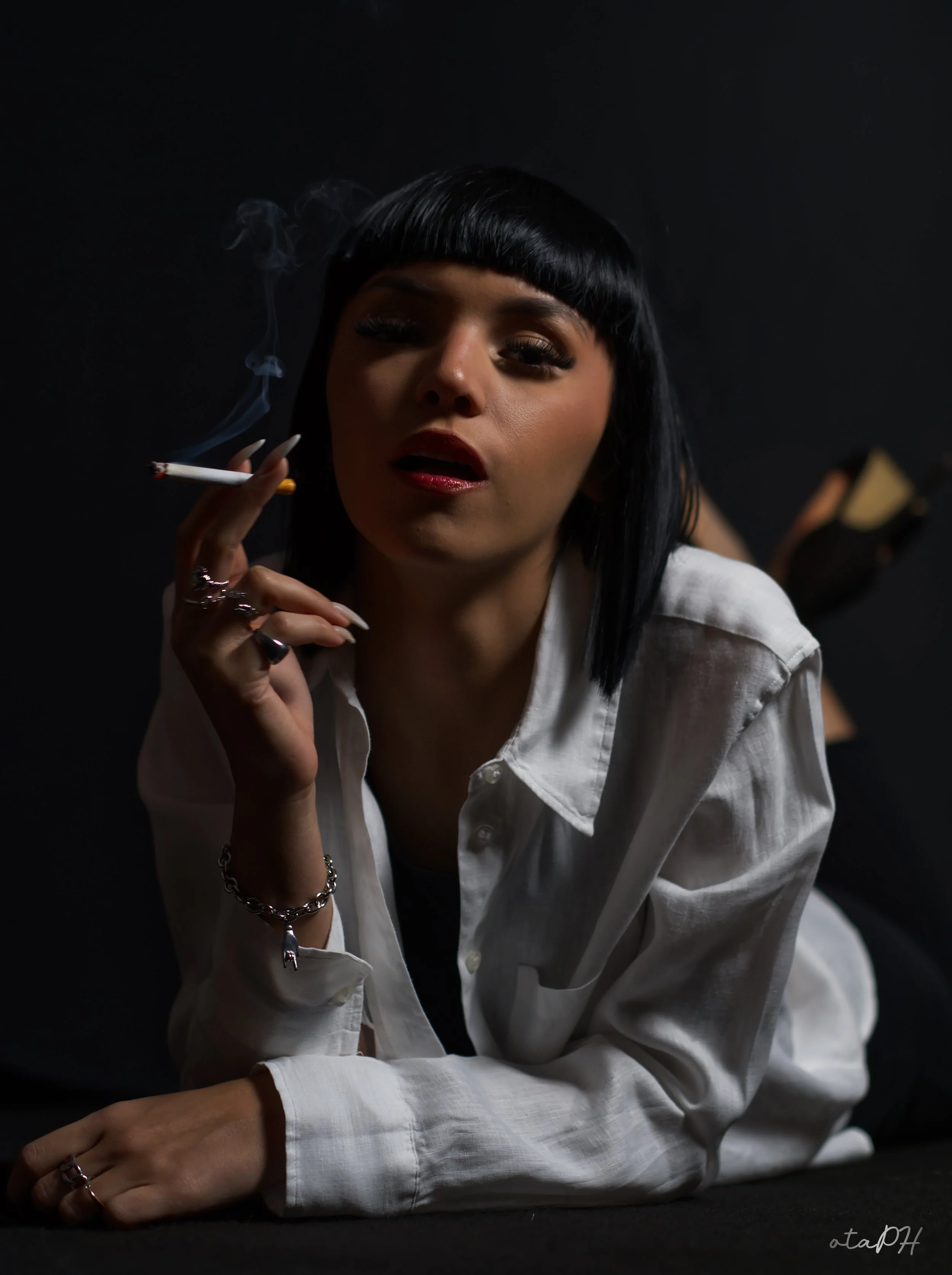 Una donna dai capelli corti e neri, con trucco intenso, indossa una camicia bianca e si regge il viso con una mano, in un ambiente scuro con sfondo nero, con sigaretta accesa e fumo visibile.
