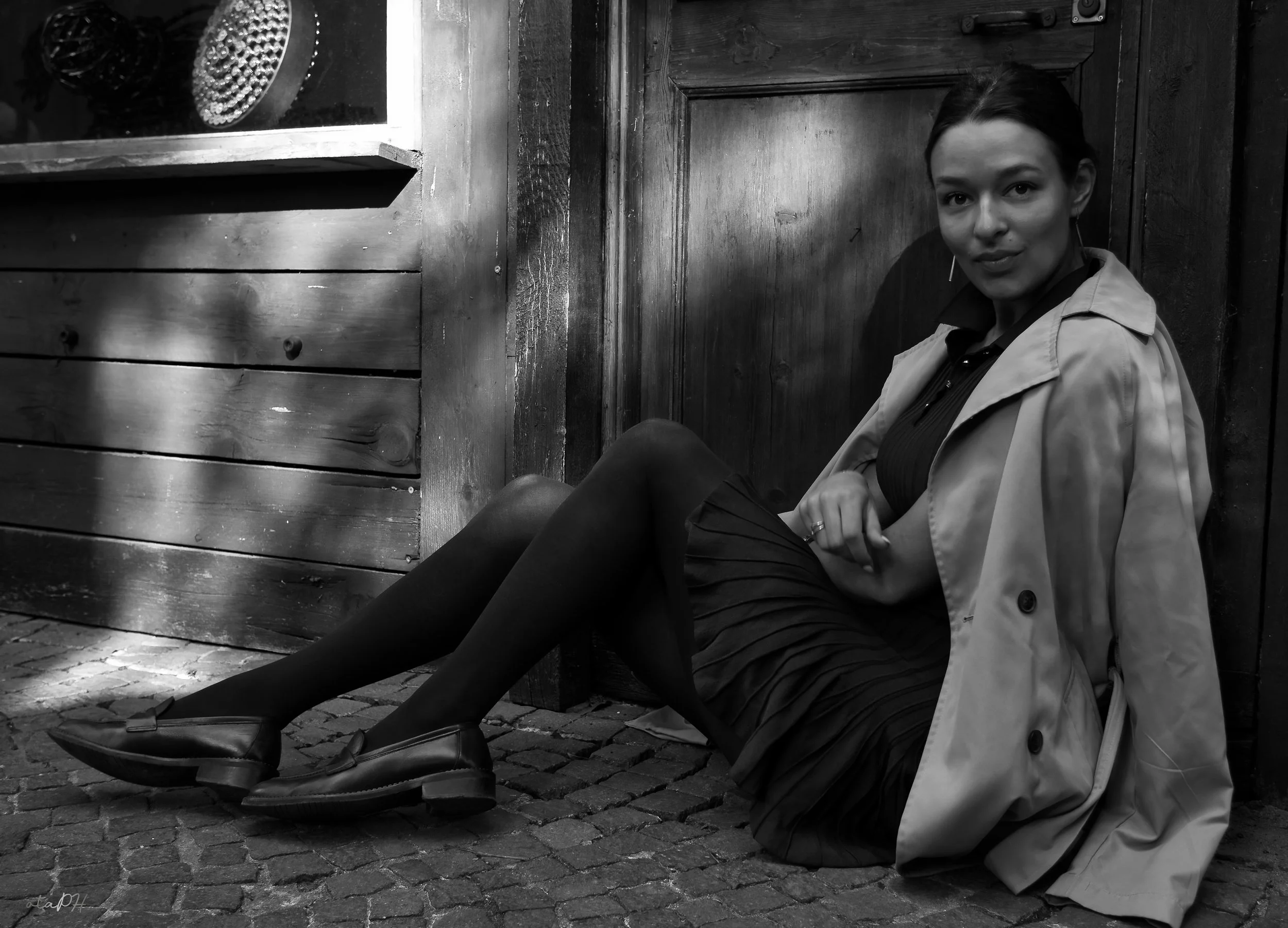 Una donna seduta per terra vicino a un muro di legno, portando un trench beige e scarpe nere con tacchi. La foto è in bianco e nero.