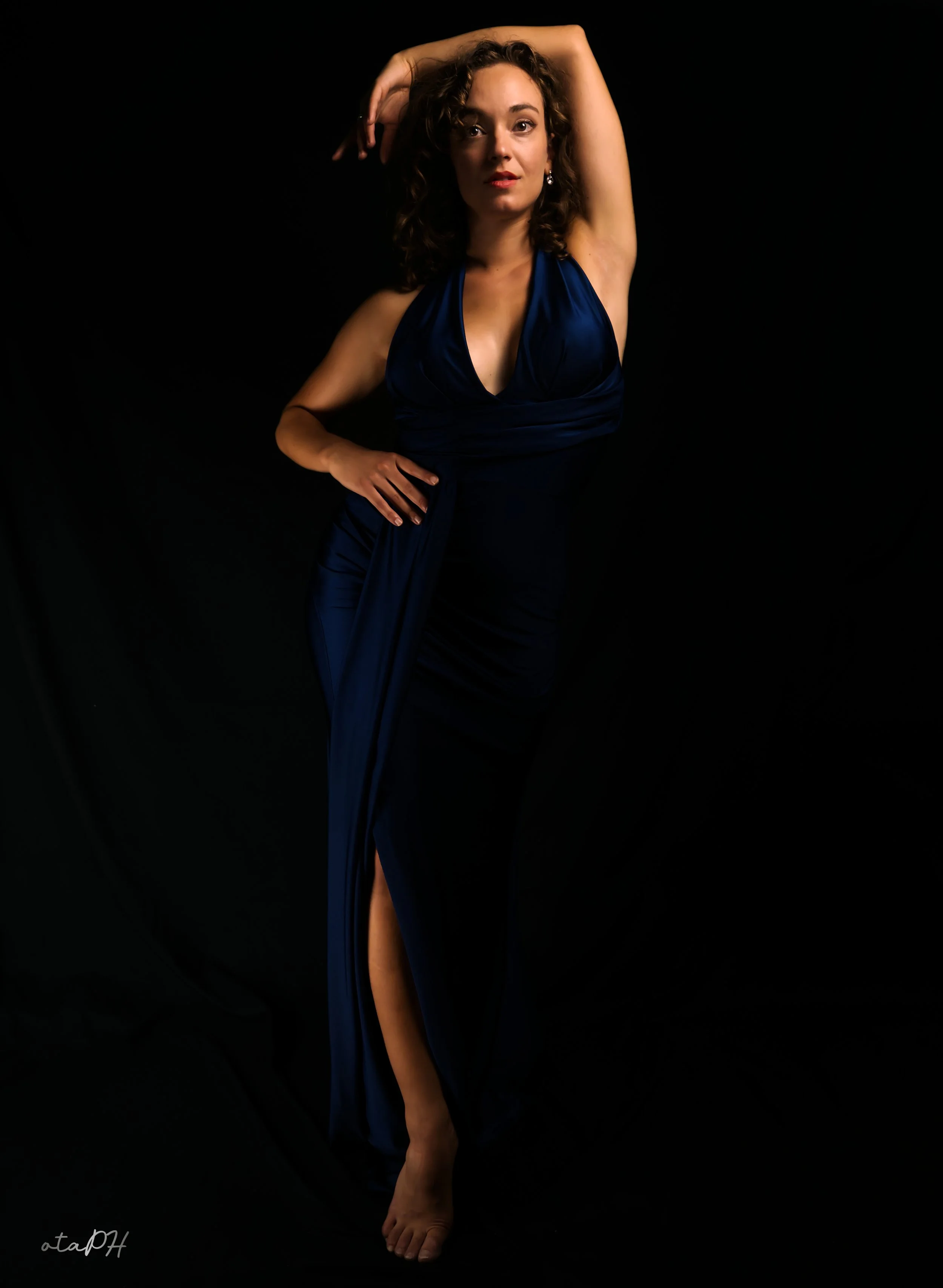 Una donna con capelli riccioli e pelle chiara indossa un vestito blu scuro, posando con una mano sulla vita e l'altra sopra la testa, contro uno sfondo nero.