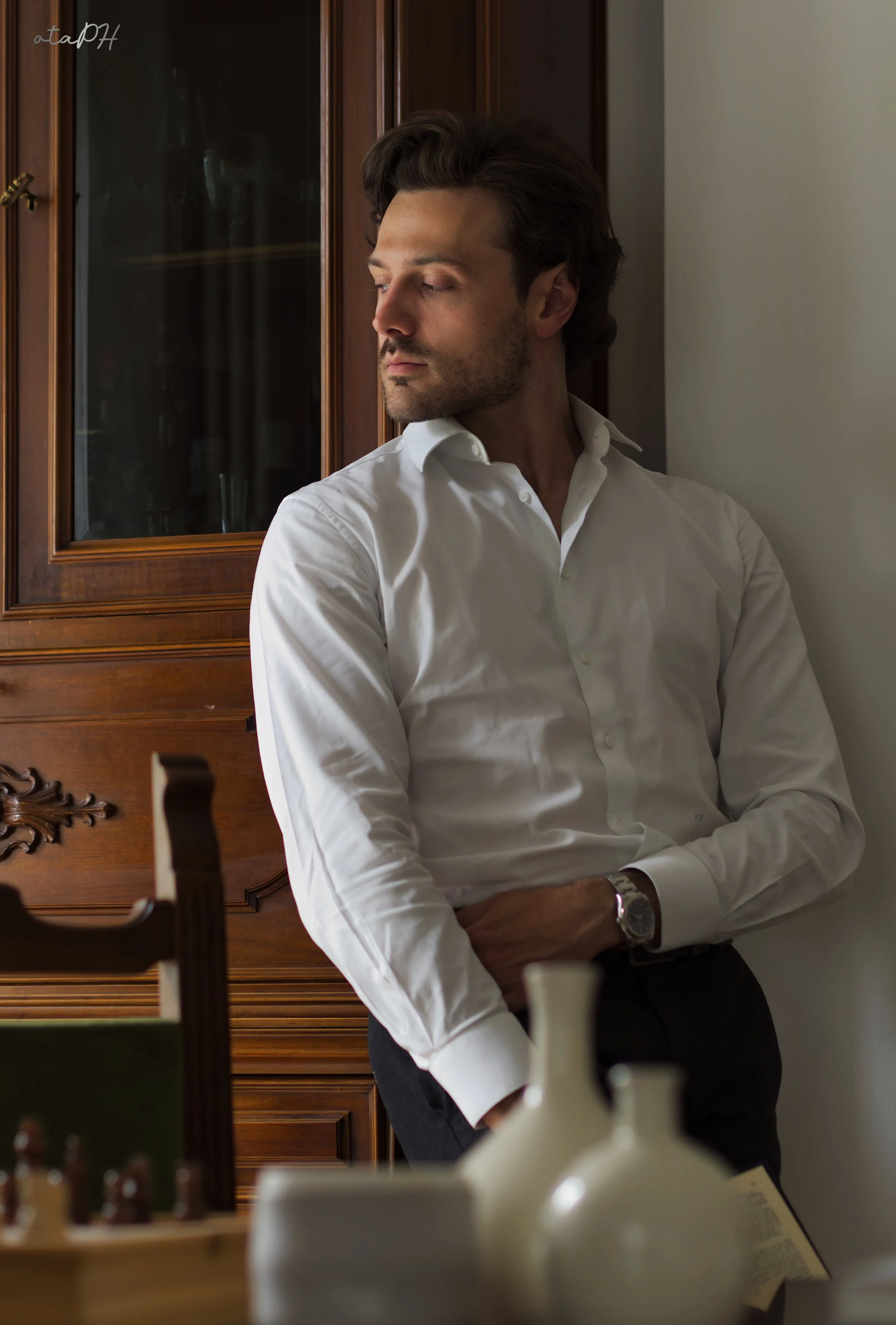 Uomo in camicia bianca con cravatta nera appoggiato a una parete, in un ambiente con mobili in legno.