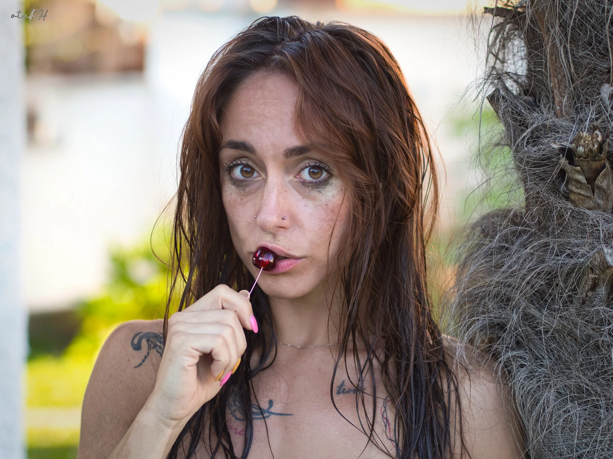 Una donna con capelli castani e rossi tiene un cioccolatino su uno stuzzicadenti vicino alla bocca, con uno sfondo di natura e una porzione di albero con cortecce e radici a destra.