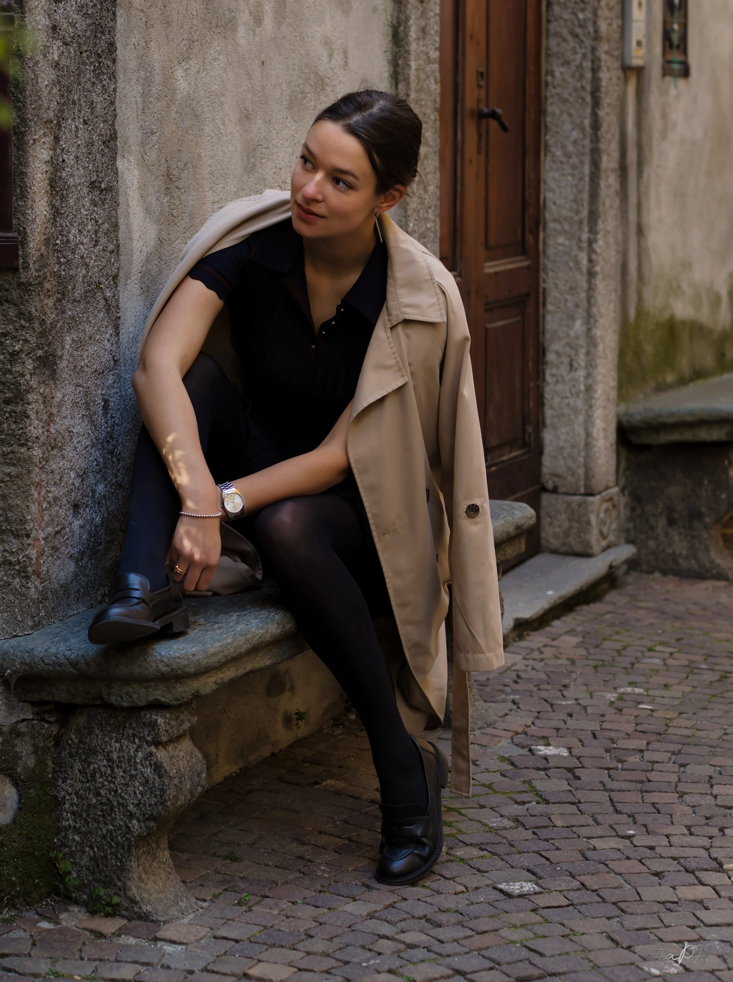 una donna seduta su un muretto in una strada di ciottoli con una giacca beige sulle spalle, indossa abbigliamento nero, un orologio e un orecchino a pendente, sfondo di pareti in cemento e una porta in legno.