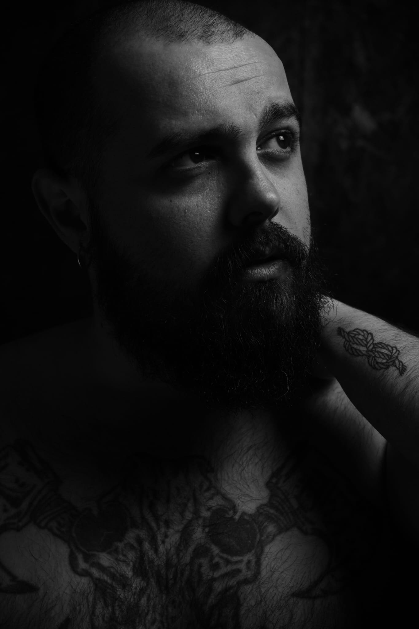 Ritratto in bianco e nero di un uomo con barba e tatuaggi, in una posa pensierosa, con uno sguardo rivolto verso l'alto e un'espressione contemplativa.