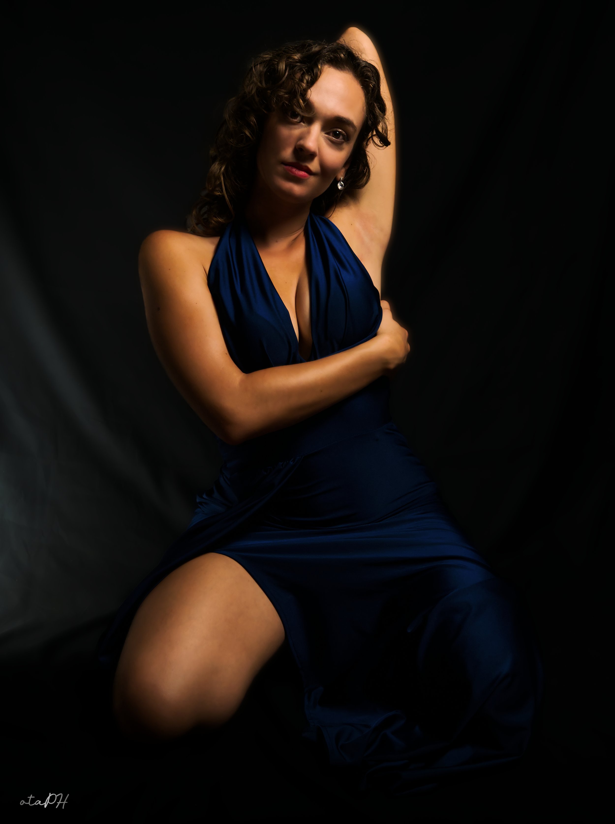 Una donna con capelli ricci, indossa un vestito blu scivolato, seduta con una gamba piegata, sfondo nero, posa con un braccio sopra la testa e uno sulle ginocchia, guarda verso la telecamera