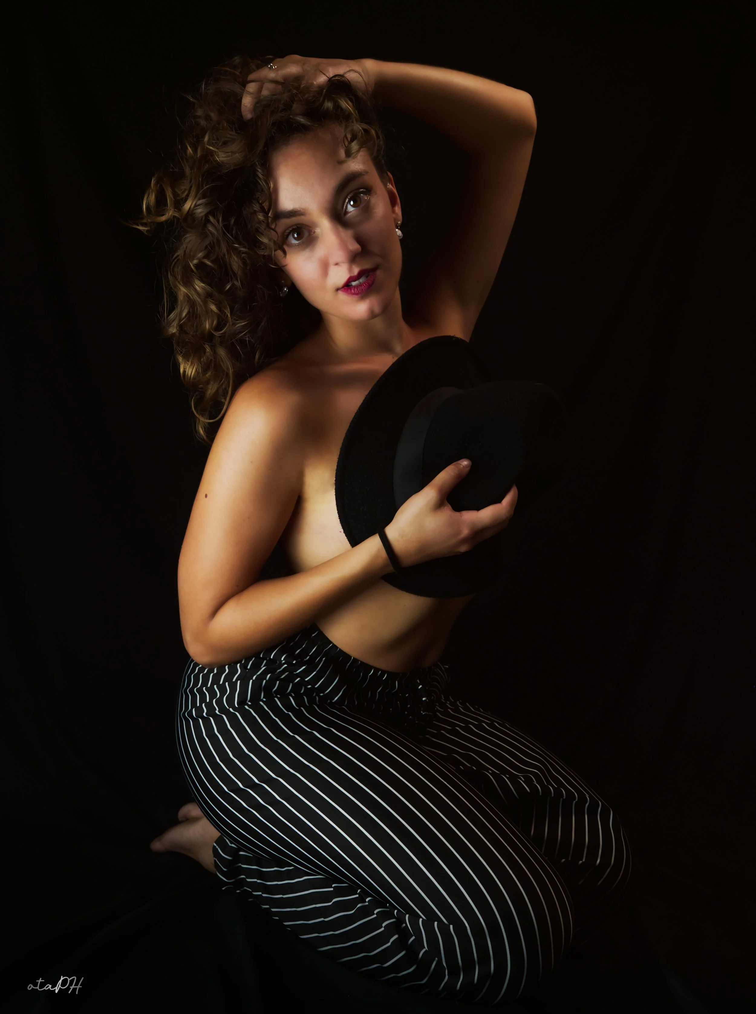 Ragazza con capelli riccioli, in posa con una mano sulla testa e l'altra che tiene un cappello nero, indossa pantaloni a righe e scrive 'otaDH' in basso a sinistra.