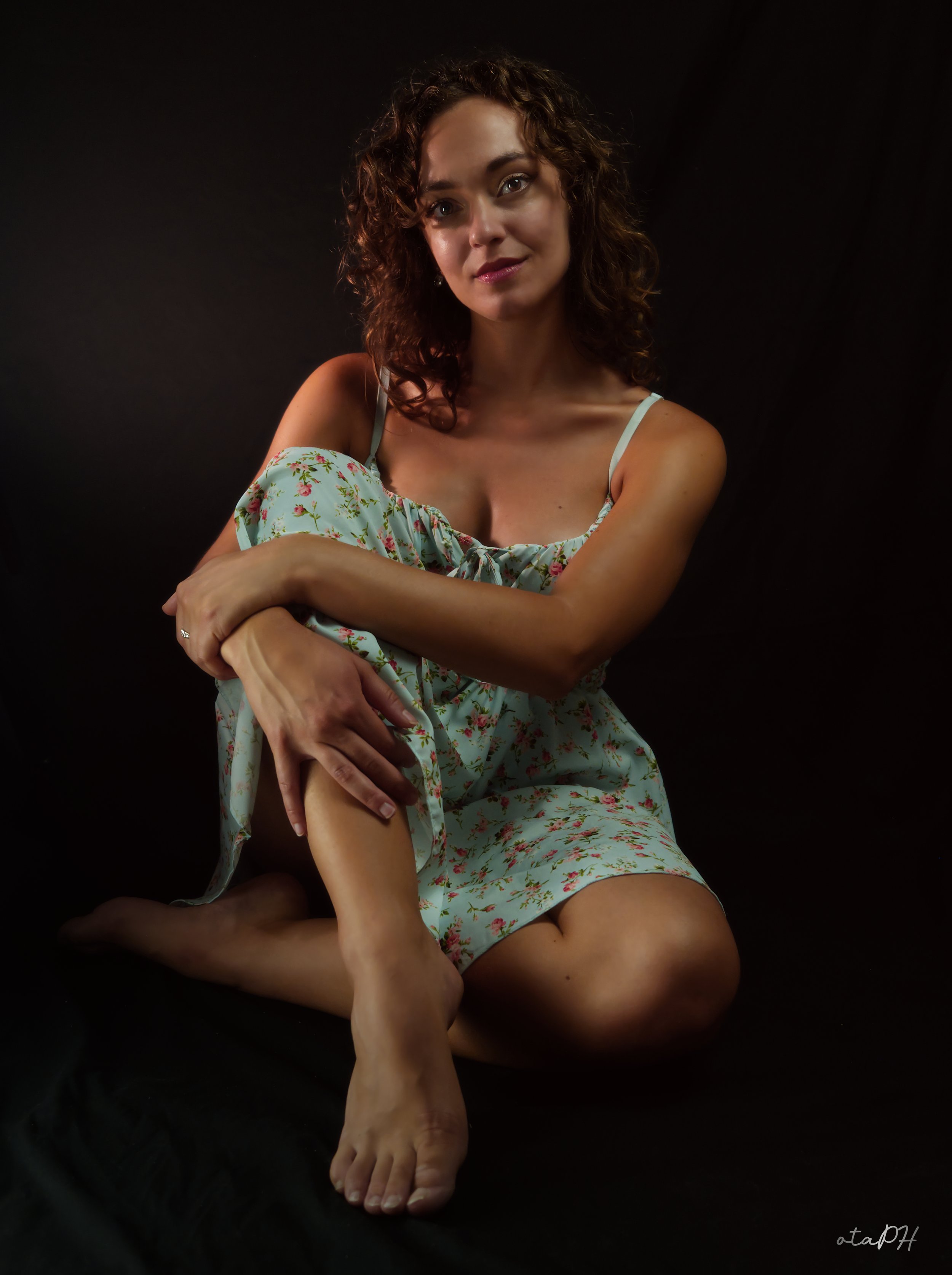 Una donna con capelli riccioli indossa un vestito a fiori con spalline sottili, seduta con le gambe incrociate, e abbraccia una gamba mentre guarda verso la telecamera con un'espressione serena. Lo sfondo è nero.