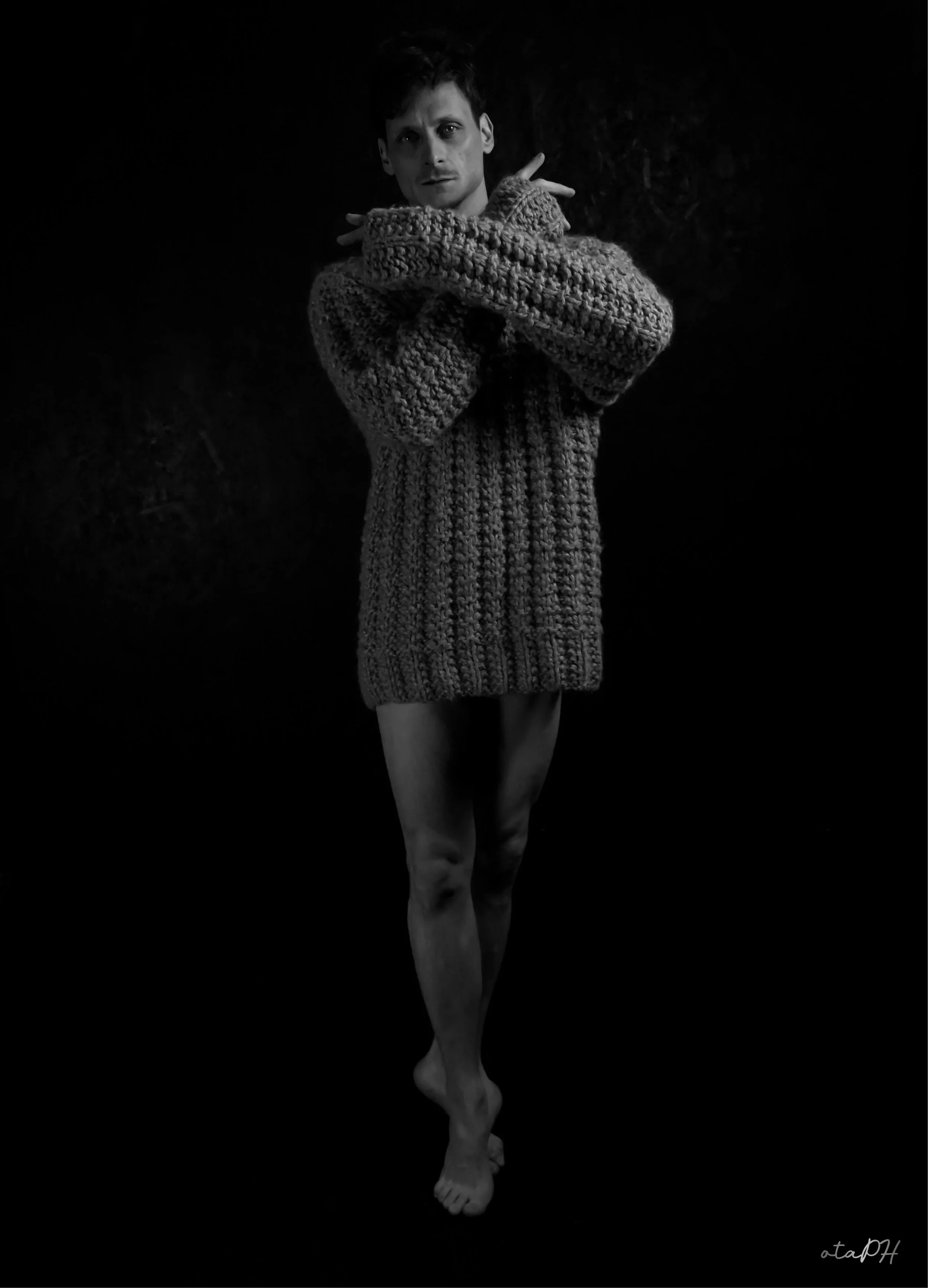 Una persona vestita con un maglione lavorato a maglia grigio chiuso davanti, con le braccia incrociate e le mani sopra le spalle, posa in uno sfondo scuro.