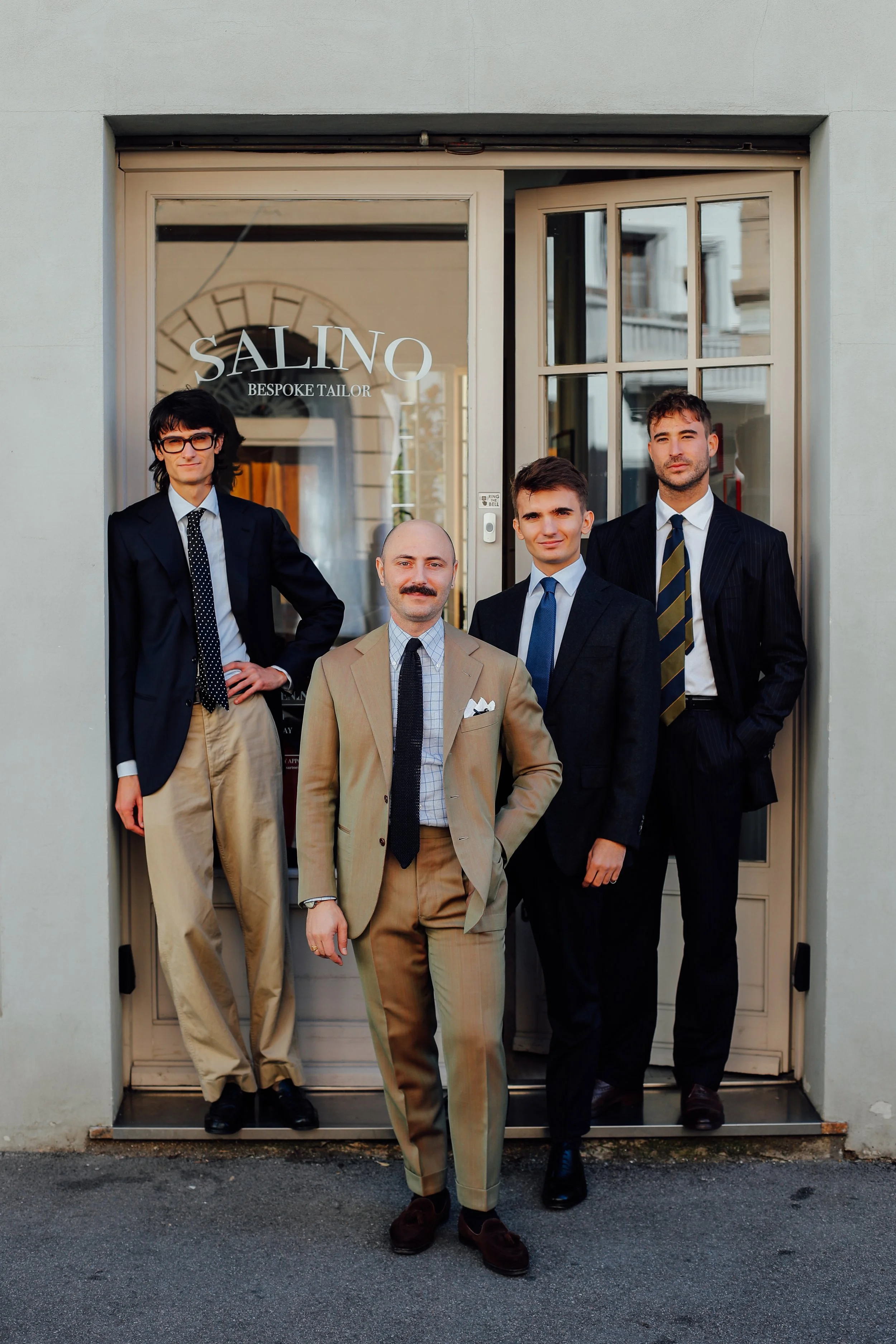 Four men in suits standing in front of a tailor shop named "SALINO BESPOKE TAILOR"; one man in beige, one in navy, and two in black suits, all dressed professionally.