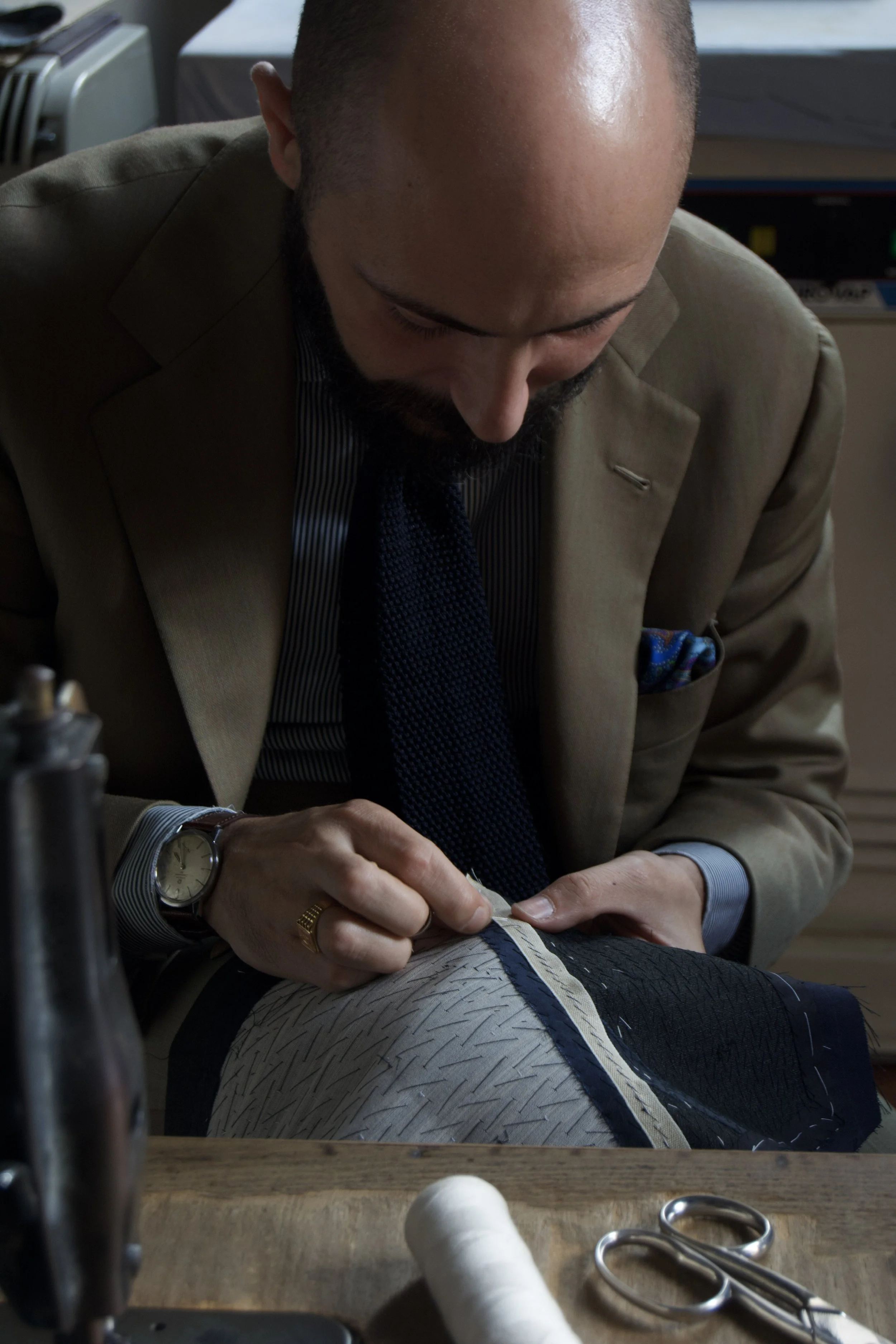 Man in a beige suit and dark tie sewing fabric at a worktable, surrounded by sewing tools and supplies.