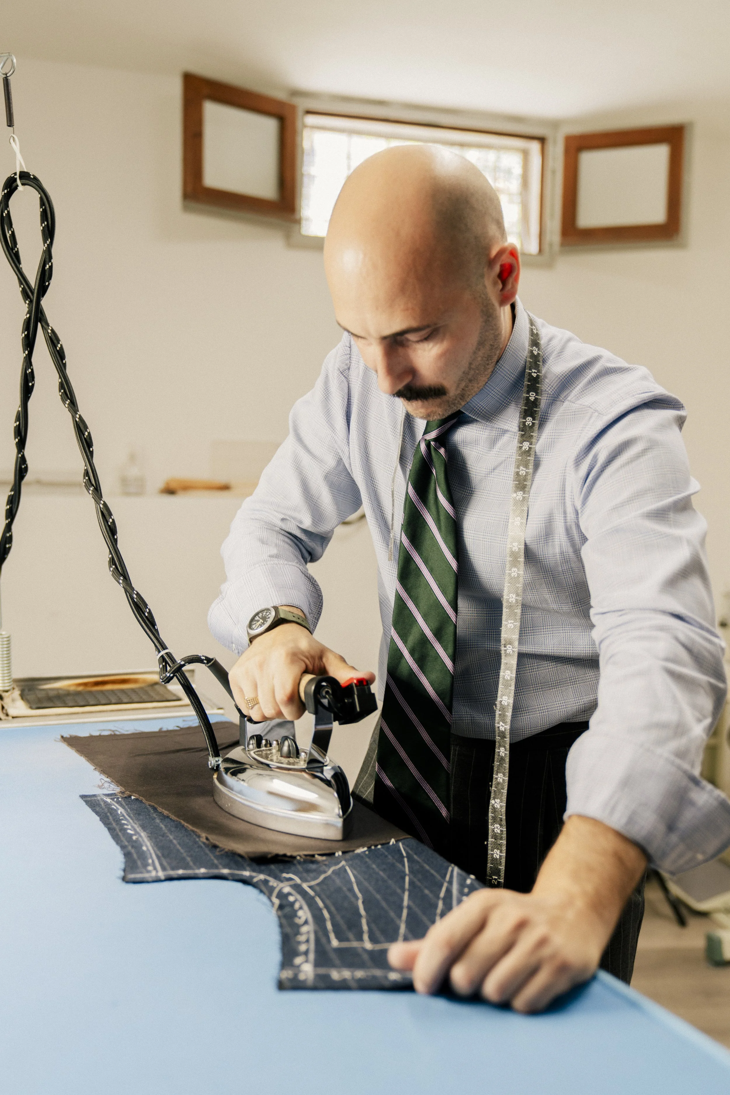 A man in a light blue dress shirt with a dark striped tie irons fabric on a blue table, next to a piece of patterned fabric, in a well-lit room with small window.