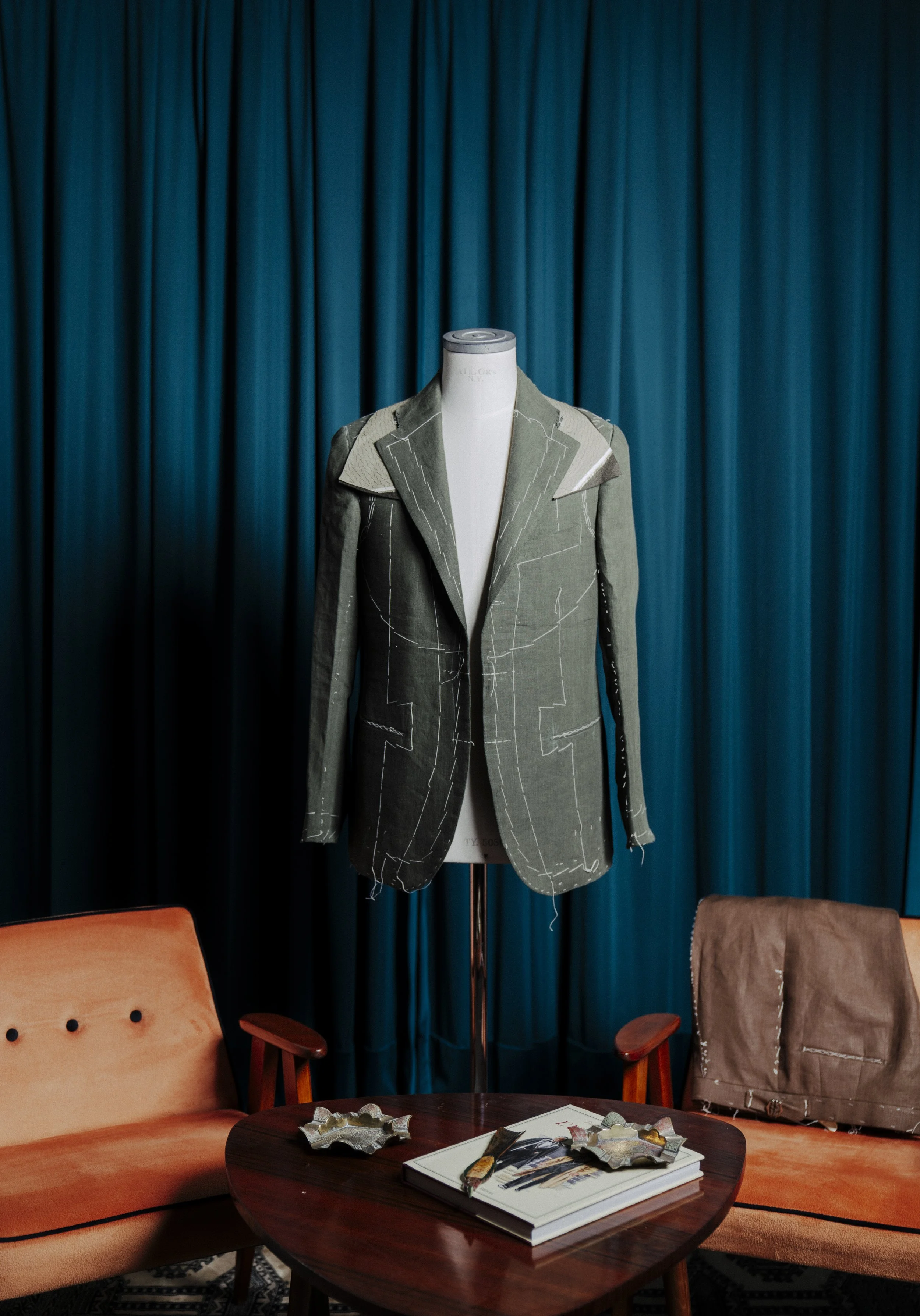 A fashion mannequin dressed in a tailored green coat with white stitching lines, placed in front of closed blue curtains. In front of the mannequin, there is a round wooden table with magazines and decorative silver coasters. Two chairs, one with an 