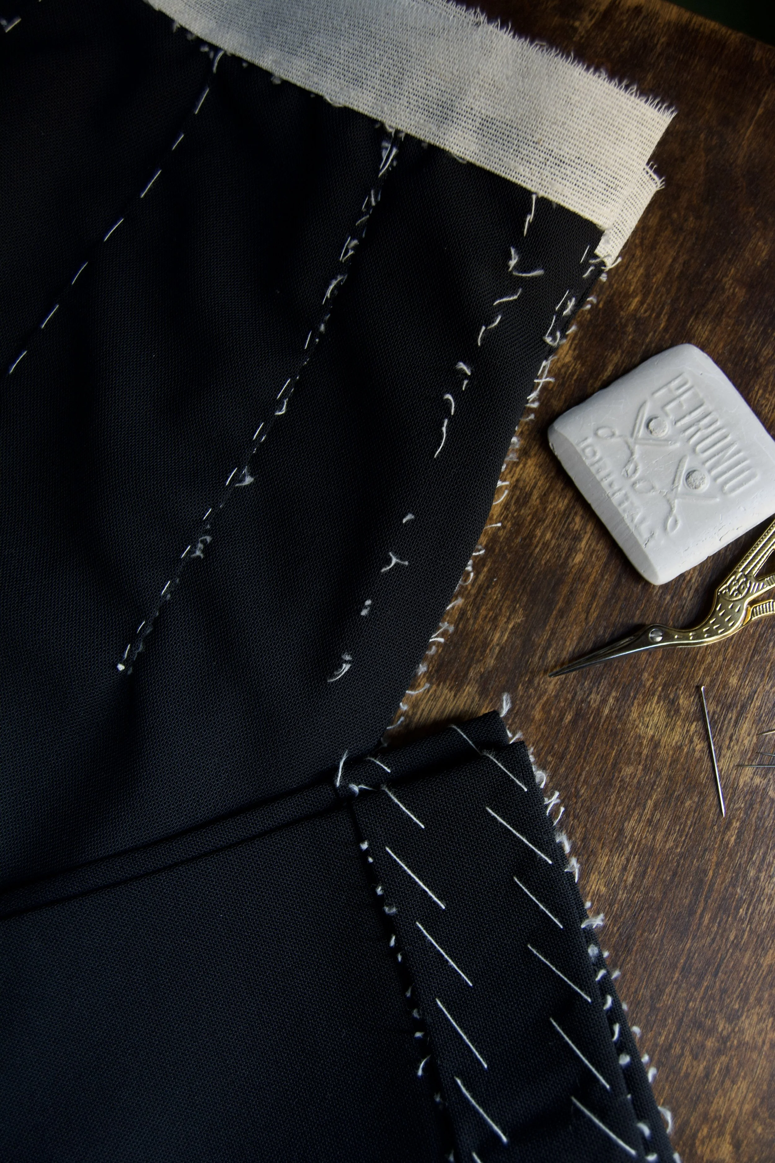 Close-up of black fabric with white stitching, sewing pins, and a white chalk stick on a wooden surface, used for sewing or tailoring.