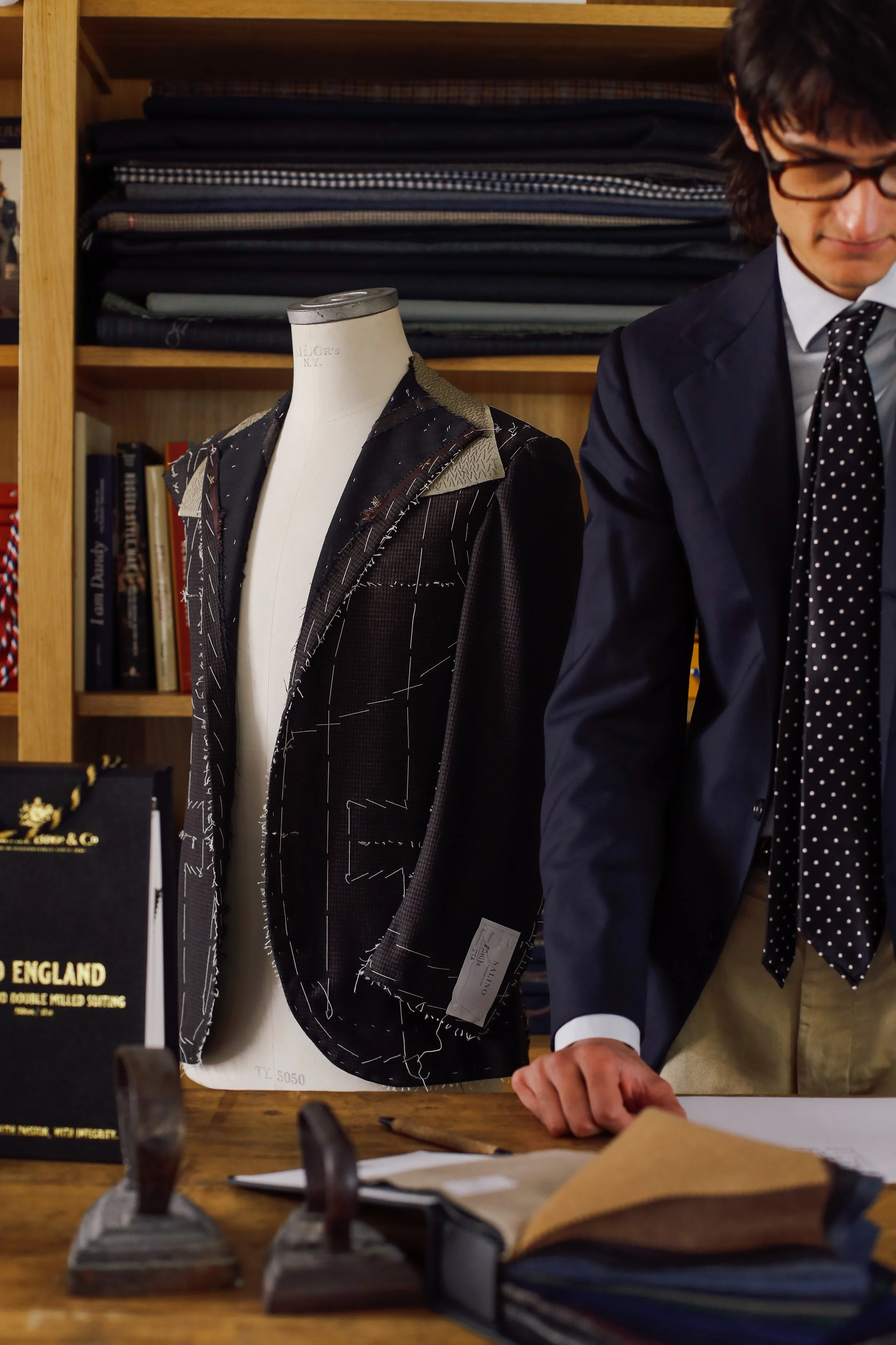A man in a suit and glasses standing in front of a tailor's dress form and shelves of fabric, with fabric swatches and books visible in the background.