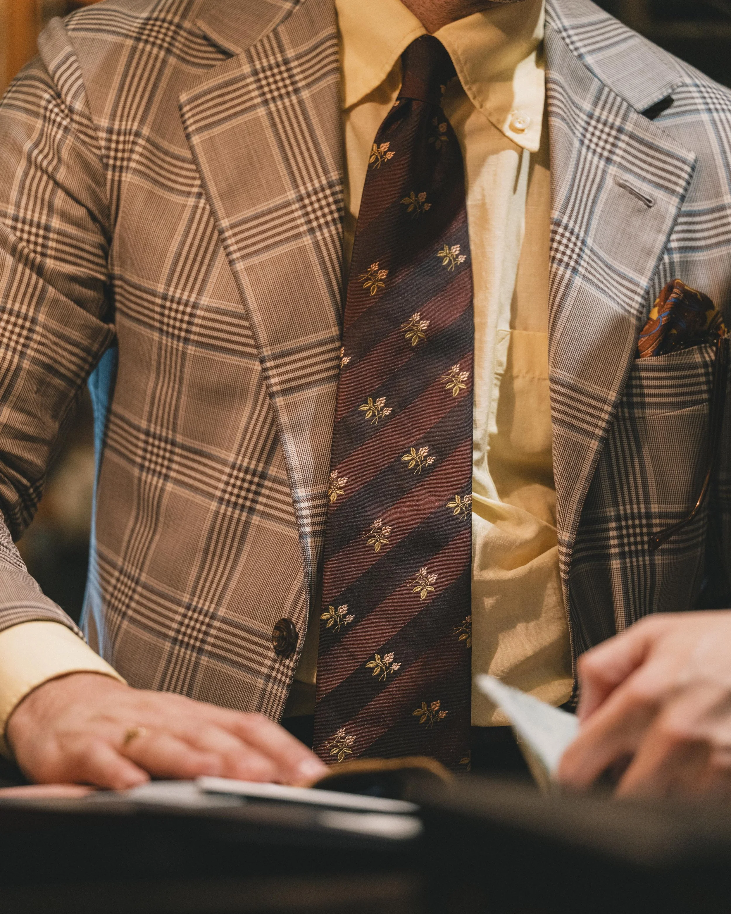 Close-up of a man wearing a beige plaid suit with a yellow shirt, patterned tie, and a pocket square, writing on a piece of paper.