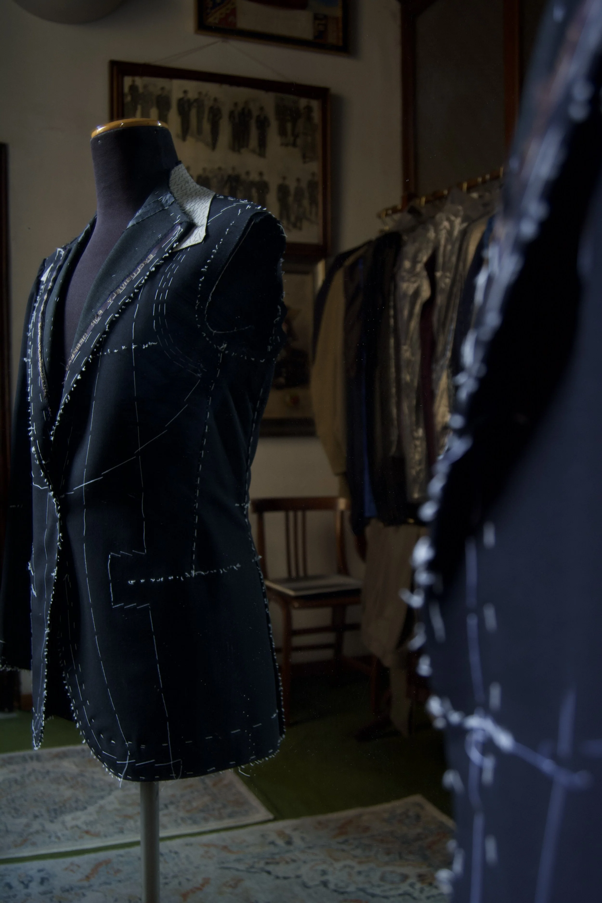 A mannequin dressed in a tailored jacket with white stitching details, situated in a room with framed photographs and clothing racks in the background.